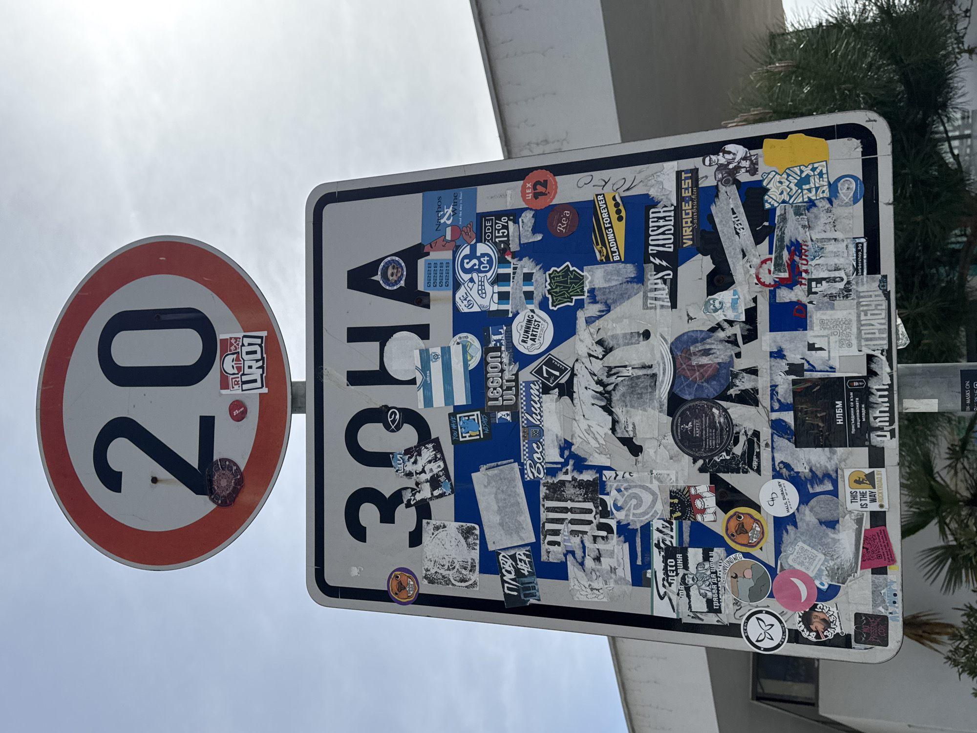 The image shows two traffic signs in Varna, Bulgaria. On the left is a circular speed limit sign indicating 20 kilometers per hour. Next to it is a rectangular sign with the Cyrillic word "ЗОНА" (ZONA), meaning "zone". Both signs are covered with numerous stickers, obscuring their original appearance. The presence of the word "ЗОНА" suggests a designated area with specific regulations. The stickers on the sign reflect a mix of commercial advertisements, local interests, and potentially political or social statements. The background includes a building facade and some greenery, suggesting an urban street setting. The presence of Bulgarian text suggests the location is in Bulgaria. The word "VARNA" is visible in the cropped image indicating the city.