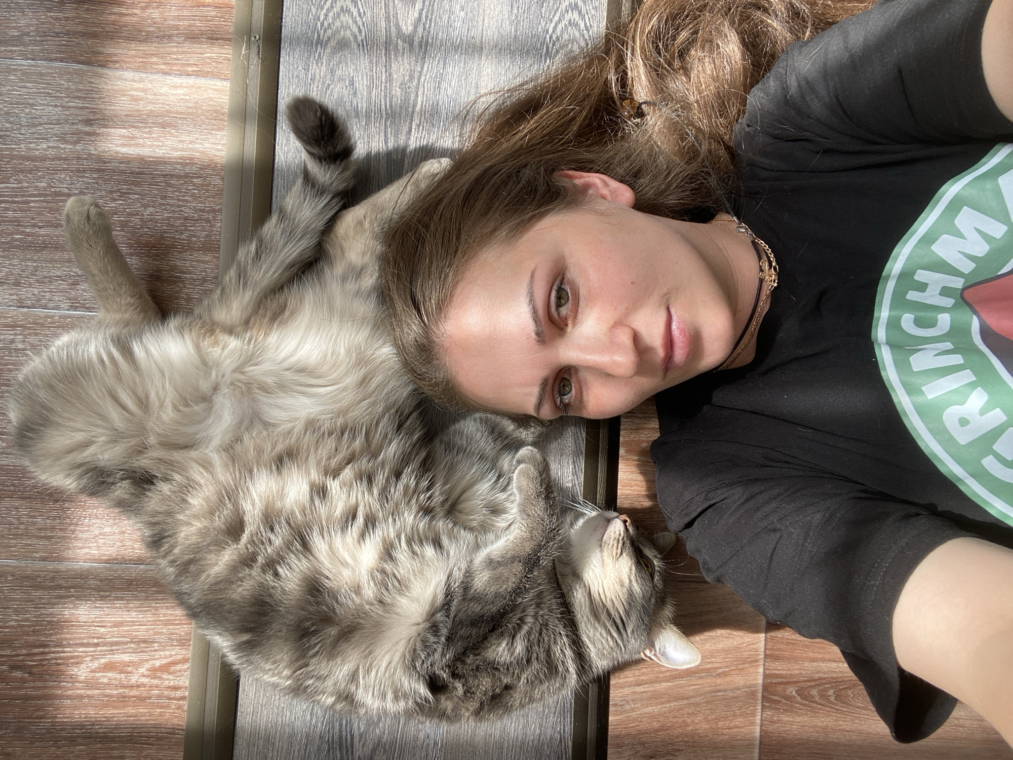 A young woman with long, light brown hair and green eyes is lying on her back on a wooden floor, basking in sunlight. She is wearing a black t-shirt with a green circular logo that reads "Grinchmas." Next to her, also on the floor, is a large, fluffy grey tabby cat lying on its back. The cat's head is turned towards the woman, and its eyes are open. Sunlight casts striped shadows across the floor and the subjects. The environment appears to be indoors, possibly a room with a wooden floor. No specific location within Марковский, Russia is discernible.