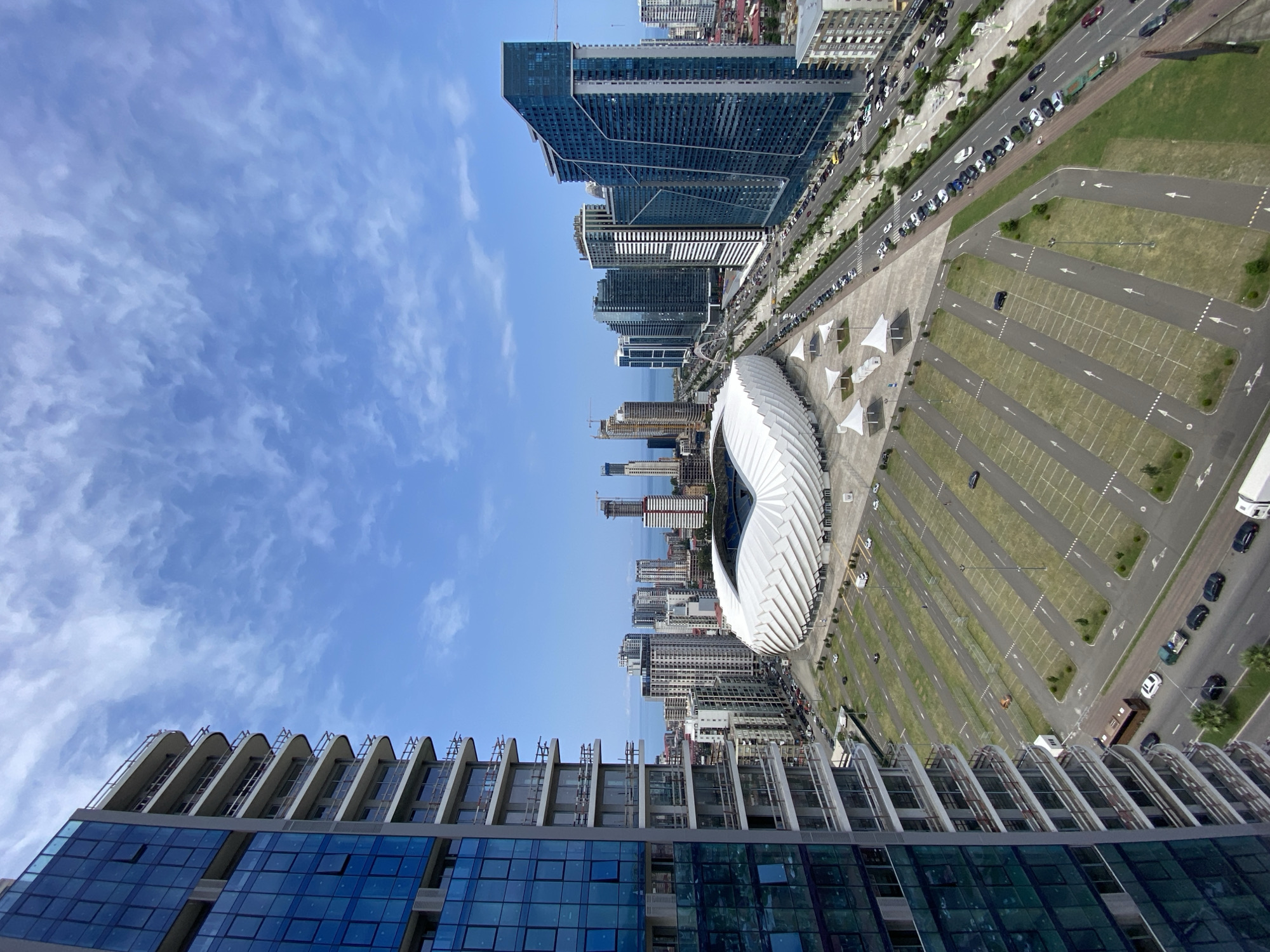 The image shows an aerial view of a modern city with a stadium, high-rise buildings, and a coastal view. In the foreground, a tall, unfinished residential building with blue glass windows is partially visible on the left. To its right, a large, white, wave-shaped stadium is prominent, with its intricate architectural design clearly visible. In the background, a dense cluster of high-rise buildings, typical of a coastal city, stretches along the horizon, with the blue expanse of the sea visible beyond them. Several roads are visible, with cars driving and parked, indicating an urban environment. The sky is a clear blue with scattered white clouds. The scene is likely the city of Batumi, Georgia, given the distinctive architecture of the stadium (Batumi Stadium) and the characteristic urban development seen along the Black Sea coast. There are no people clearly visible in the image.