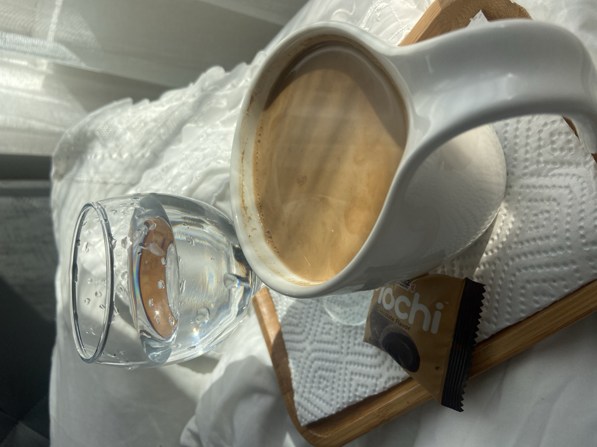 The image depicts a close-up still life scene, seemingly on a white, textured bedspread. A white ceramic mug filled with a light brown beverage, possibly coffee with milk, rests on a small wooden tray lined with a white napkin. Next to the mug is a small package of "Mochi" Chocolate Flavor snack. A clear glass containing a golden liquid sits adjacent to the tray. The scene is bathed in natural light, possibly from a nearby window. The image provides no visual indication of location.