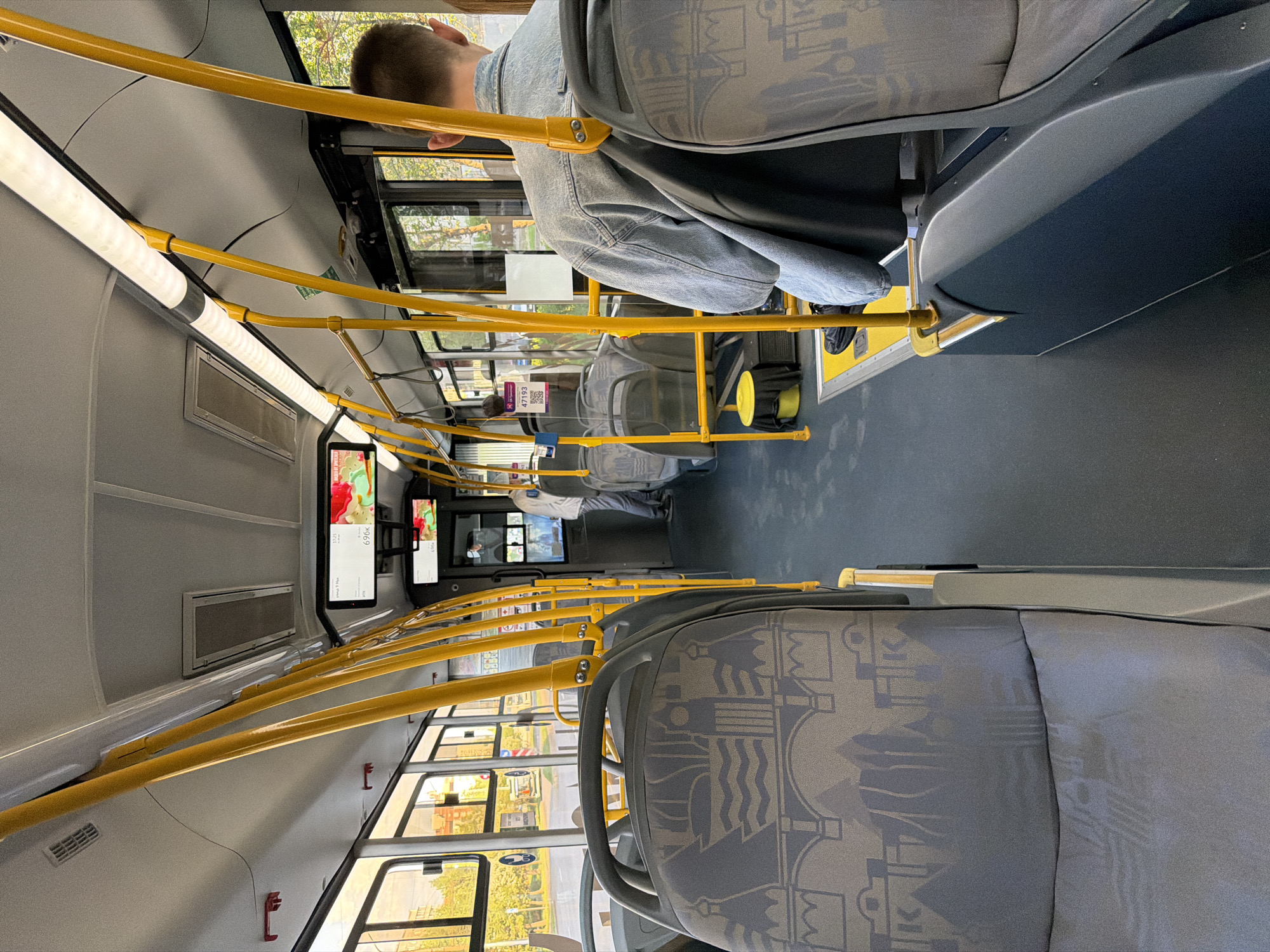 The image shows the interior of a bus, seemingly in Krasnoyarsk, Russia. The interior features rows of gray patterned seats with yellow handrails. Passengers are seated, with one wearing a denim jacket and others visible further down the aisle. Digital screens display information or entertainment. Outside the windows, buildings and greenery suggest an urban or suburban setting. The bus appears modern and well-maintained.