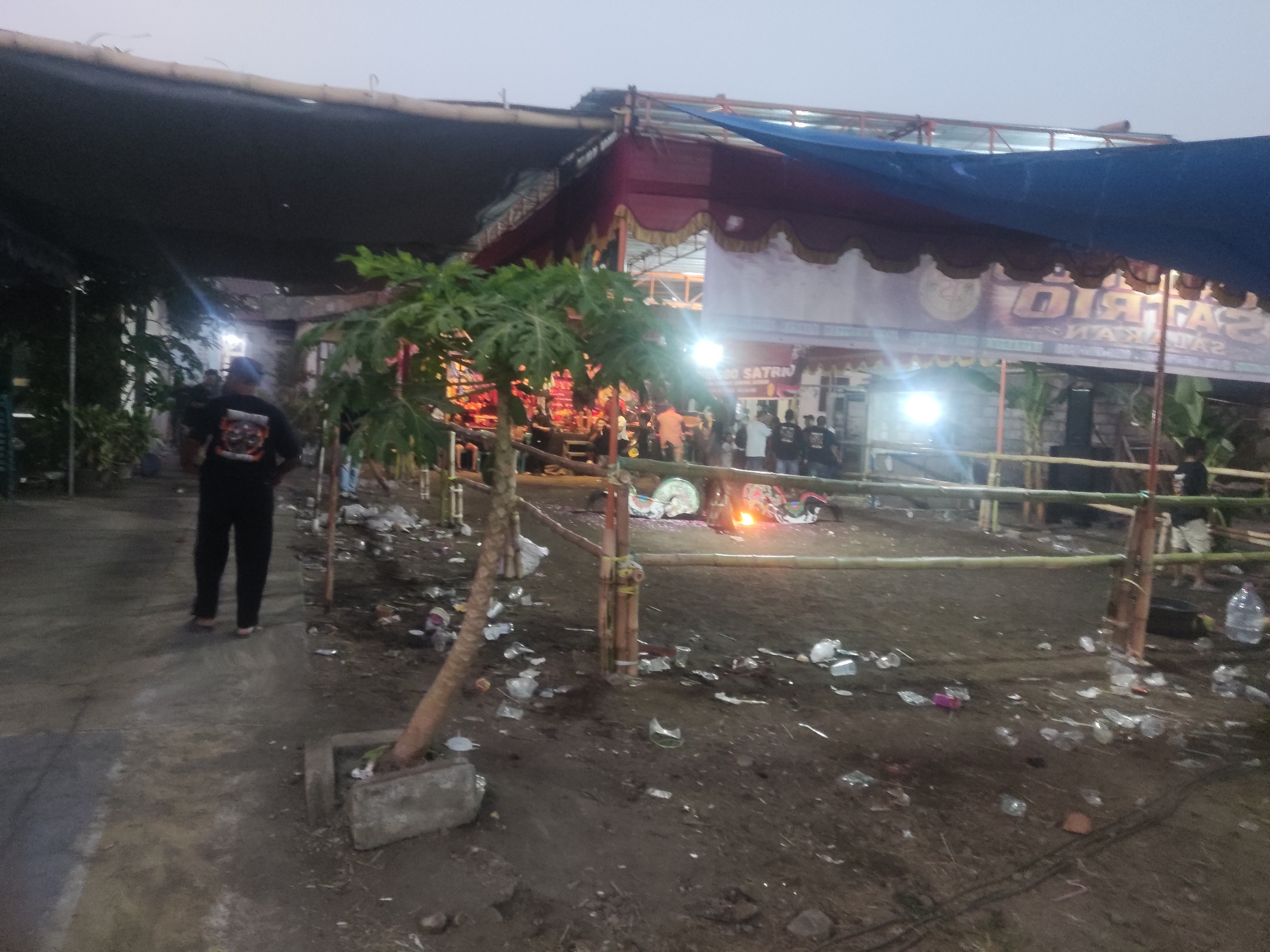 The image depicts an outdoor scene at dusk in Trihanggo, Indonesia, likely after an event. A temporary stage structure covered by a blue tarp and red fabric is visible in the background, with bright lights illuminating the area. Several people are gathered, some standing near the stage and others walking around. The ground is covered in dirt and littered with discarded plastic bottles and other debris, indicating a significant amount of waste accumulation. A small tree is planted in a concrete planter in the foreground. The overall impression is one of a post-event cleanup site with lingering signs of activity.