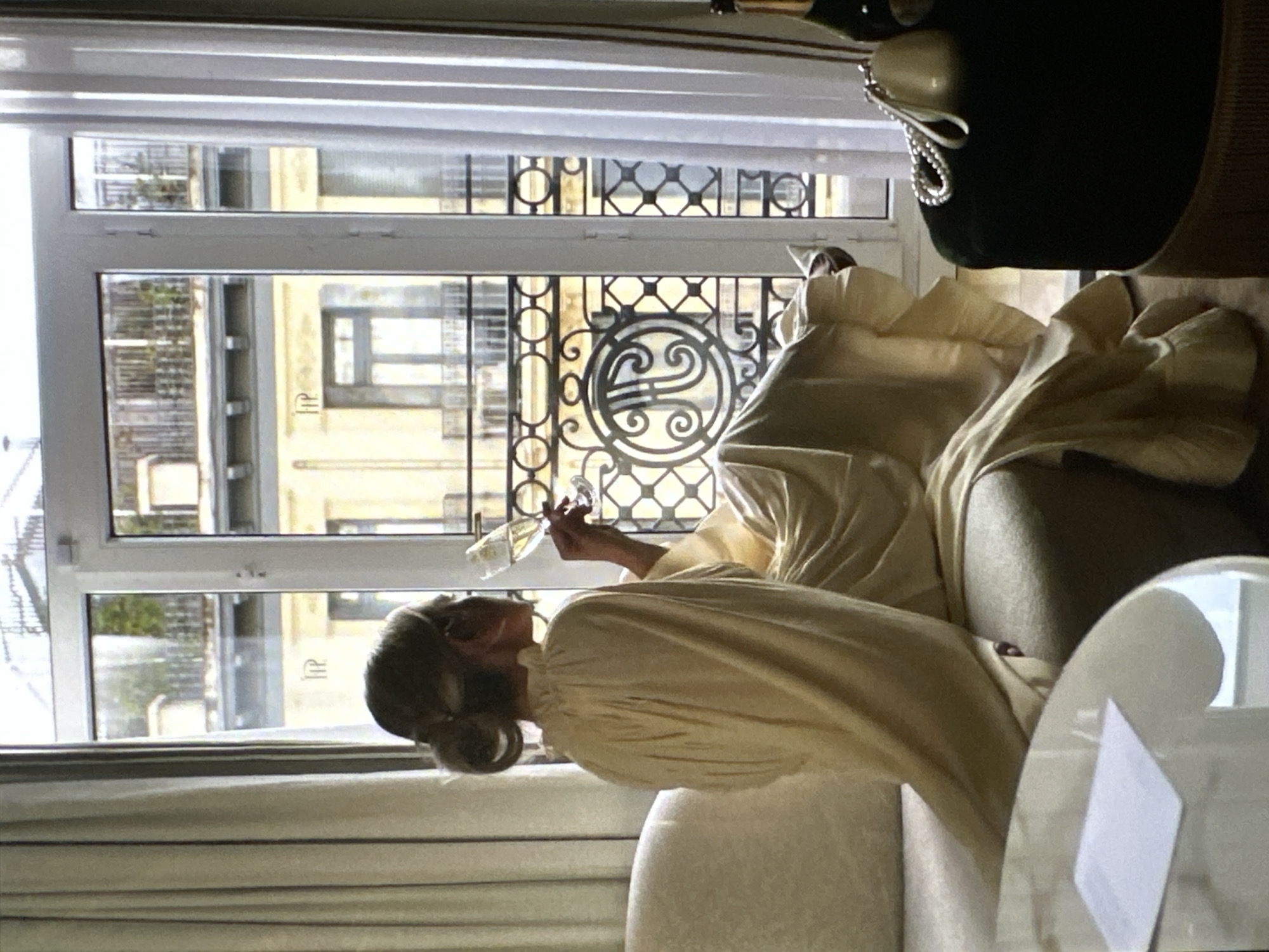 A woman in a light-yellow garment is sitting on a sofa, facing a window with ornate metalwork. She is holding a wine glass in her right hand. The building across the street features a logo that appears to be "HP". A black, velvet-covered object with pearl embellishments is partially visible in the upper right corner. The location is likely Yerevan, Armenia, based on the architectural details and signage.