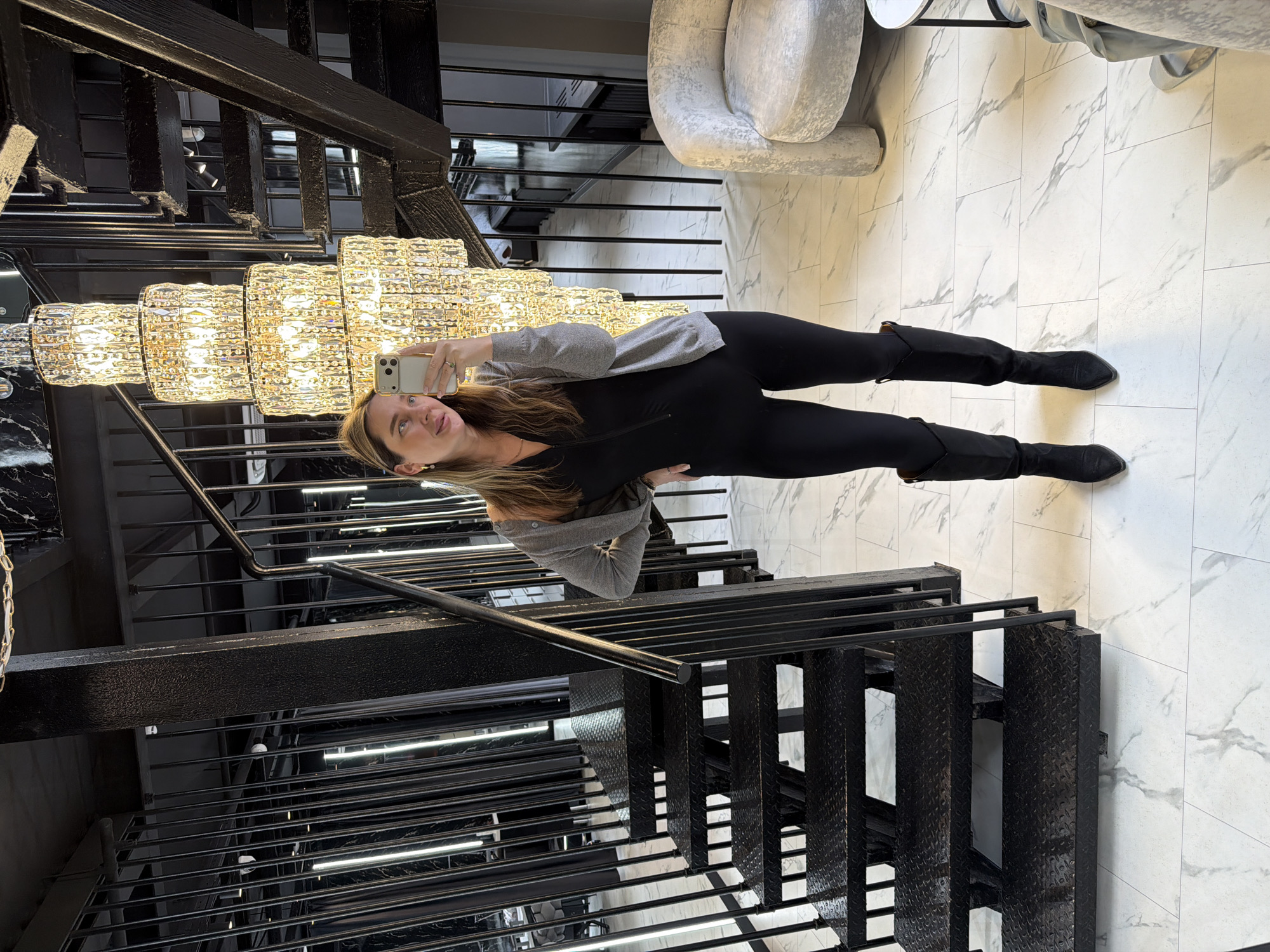 A woman with long blonde hair is taking a selfie using a white smartphone. She is wearing a grey sweater and black leggings and black boots. She is standing on a marble-tiled floor in front of a black metal staircase, underneath a large crystal chandelier. The location appears to be indoors, possibly in a building with modern architectural features. The city of Moscow, Russia is not identifiable in the image.