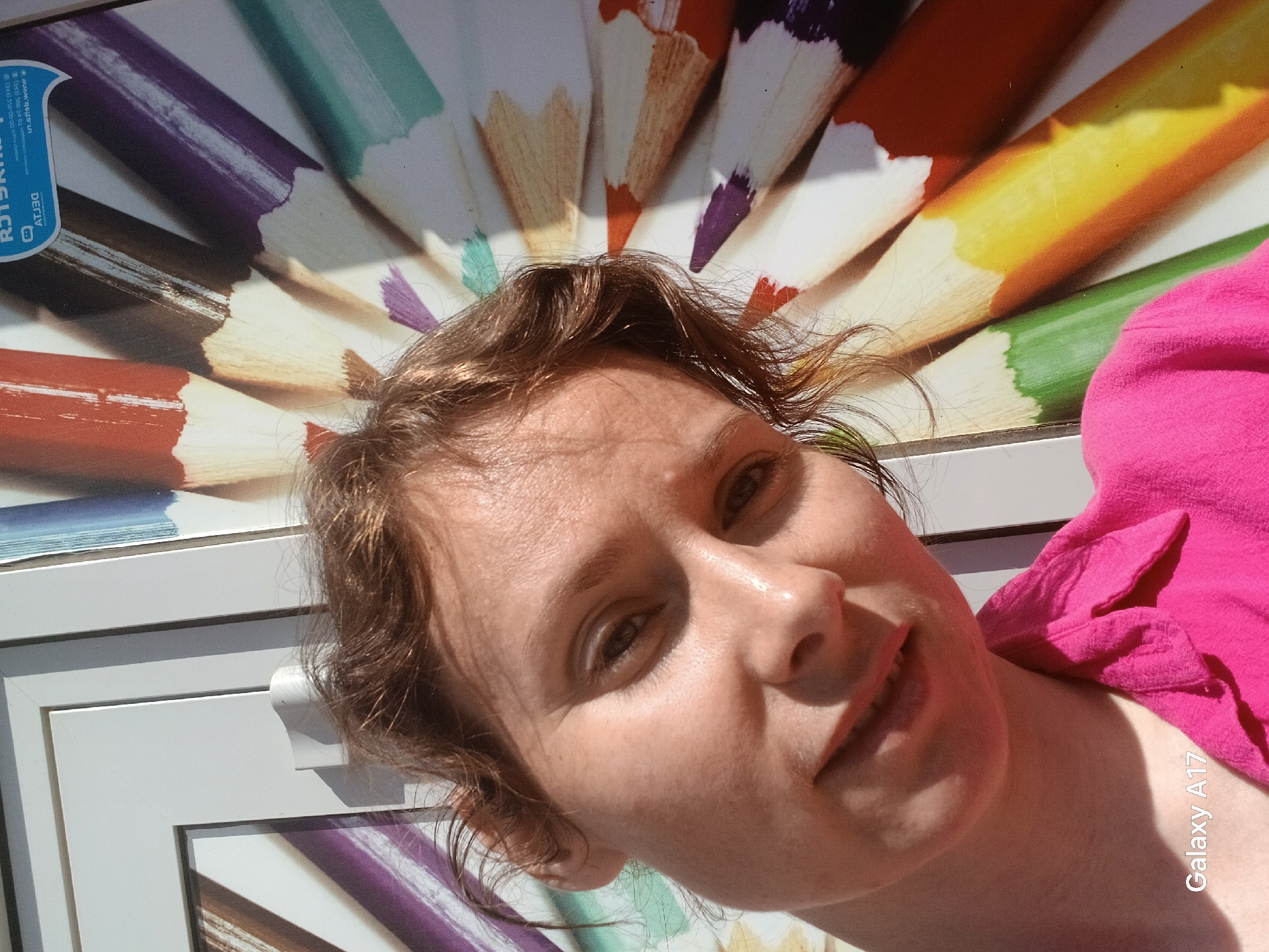 A woman is photographed indoors in Yekaterinburg, Russia. The woman has light brown hair and is wearing a bright pink top. She is positioned in front of a wall featuring a poster or display of colored pencils arranged in a semi-circular fan pattern. The pencils range in color from purple and brown to yellow and green. The frame of the artwork is visible behind and to the left of the woman. The text "Galaxy A17" is digitally added in the lower right corner of the image.