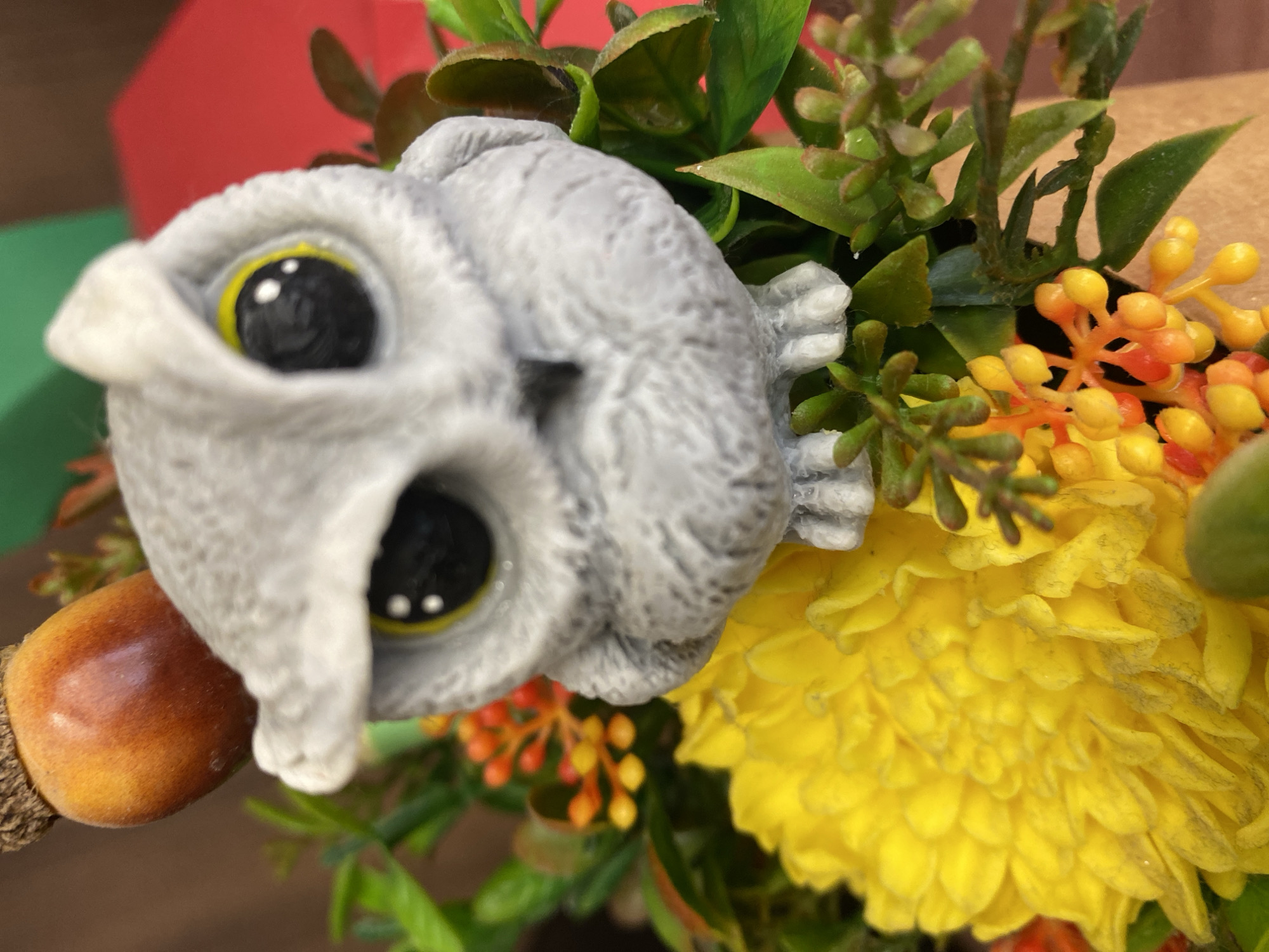 The image shows a close-up of a decorative arrangement. The focal point is a gray, owl-shaped object with large, black eyes rimmed with yellow. To the left of the owl is an acorn. To the right are greenery, small orange berry-like items, and a large yellow flower resembling a chrysanthemum or dahlia. The scene includes no people and appears to be a static still life composition, possibly indoors. There is no indication of Chaikovsky, Russia in the image.
