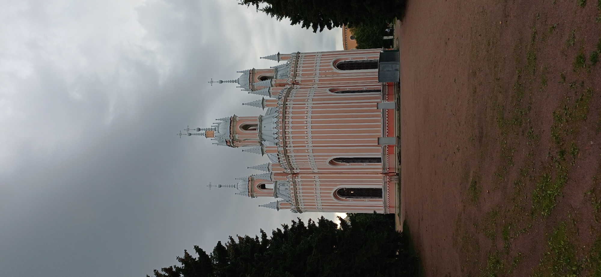 The image shows the Chesme Church in Saint Petersburg, Russia, on a cloudy day. The church is a striking example of Gothic Revival architecture, with its vibrant pink exterior and intricate white detailing. It features multiple onion domes topped with crosses, tall arched windows, and a symmetrical, octagonal design. The church is surrounded by a park-like area with sparse green vegetation on the ground and two large evergreen trees framing the structure on either side. The sky is overcast with dark, heavy clouds, suggesting an impending storm or simply a gloomy day. In the background, beyond the trees, hints of other buildings and foliage can be seen. A few indistinct figures are visible near the church entrance and in the background, suggesting the location is accessible to the public. The overall impression is one of historical grandeur set against a somber sky.