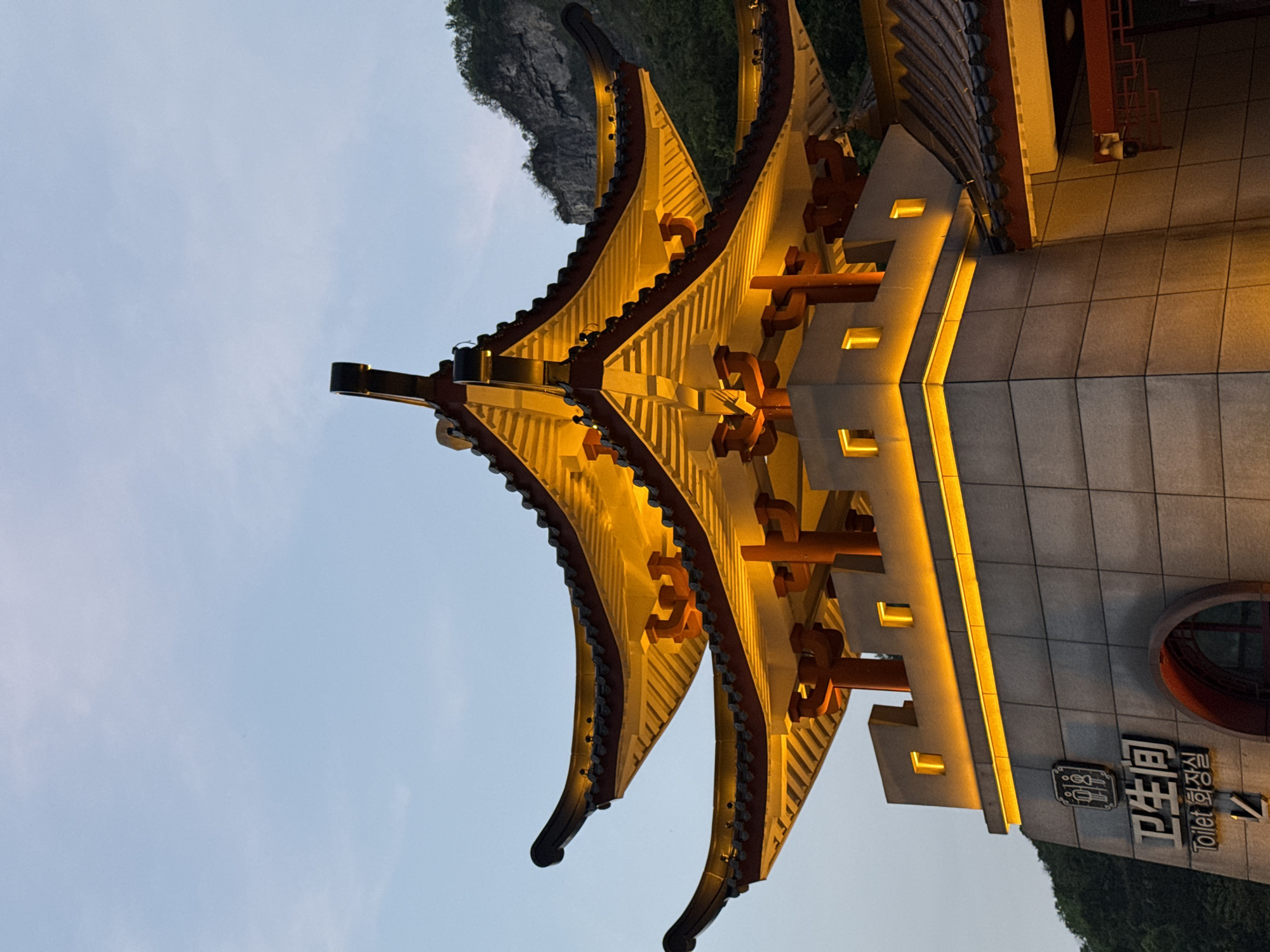 A nighttime shot shows a building with traditional Chinese architecture, specifically a public toilet sign. The scene takes place in Yongding District, China. The structure has a multi-tiered roof with ornate, upturned eaves illuminated by warm-toned lighting. The walls are tiled in shades of gray and feature strategically placed lighting accents, highlighting architectural details. Chinese characters and English text "Toilet" appear on a sign attached to the building. A glimpse of natural surroundings, including a rocky formation and greenery, are seen in the background. The sky is a light, soft blue.