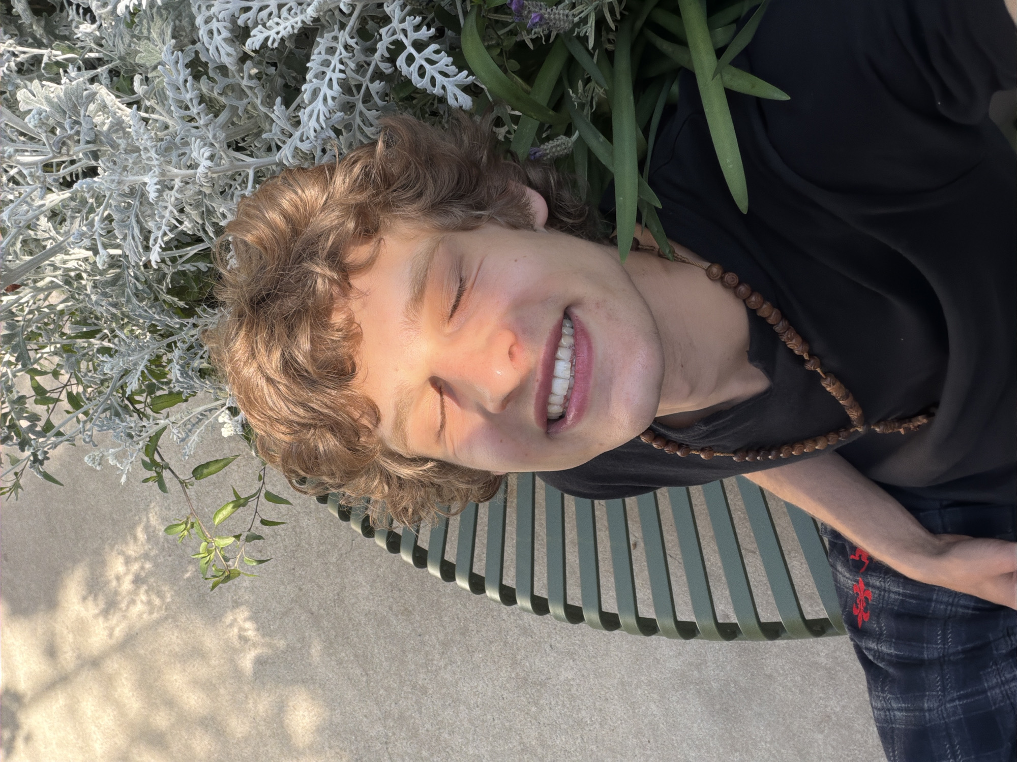The image, taken in Milan, Italy, captures a young individual relaxing outdoors under bright sunlight. The subject has light brown, curly hair and fair skin, and is seen smiling broadly with their eyes closed, conveying a feeling of serene enjoyment. They are wearing a black short-sleeved top and plaid pants, which feature a distinct red embroidered symbol or pattern. A long, wooden beaded necklace is draped around their neck.
The person appears to be nestled among lush plants. A prominent bed of silvery-green, textured foliage, possibly Dusty Miller, occupies the upper left portion of the frame and extends around their head. Darker green leaves and delicate purple flowers, resembling lavender, are also visible. The individual seems to be resting on a green slatted bench, which is partially visible towards the bottom right. Below the bench and to the left, light-colored concrete pavement is seen, marked with clear shadows, indicating strong, overhead sunlight consistent with a clear midday or early afternoon. The overall scene exudes a peaceful and relaxed atmosphere.