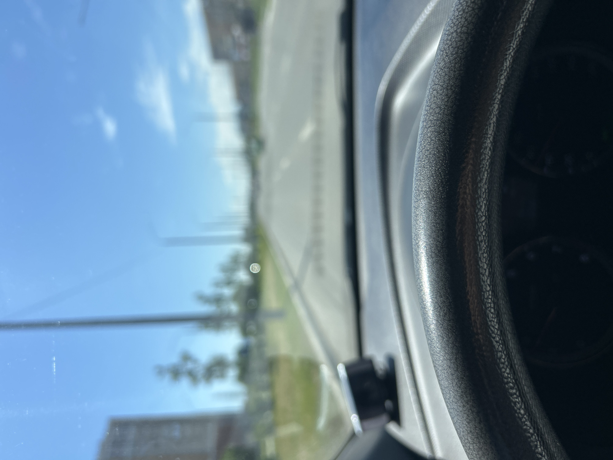 The image, taken from inside a vehicle in Orenburg, Russia, shows a view through the windshield. The foreground is dominated by a dark-colored steering wheel, which appears worn and textured. To the left of the steering wheel, on the dashboard, is a small, circular black object, possibly a dashcam or other electronic device.
Beyond the dashboard, the view opens up to a bright, sunny day with a clear blue sky dotted with a few wispy clouds. A road stretches into the distance, lined with streetlights and trees. Buildings, appearing somewhat blurred due to the focus, are visible on either side of the road, suggesting an urban or suburban environment. The overall impression is that of a car driving on a well-maintained road on a pleasant day.