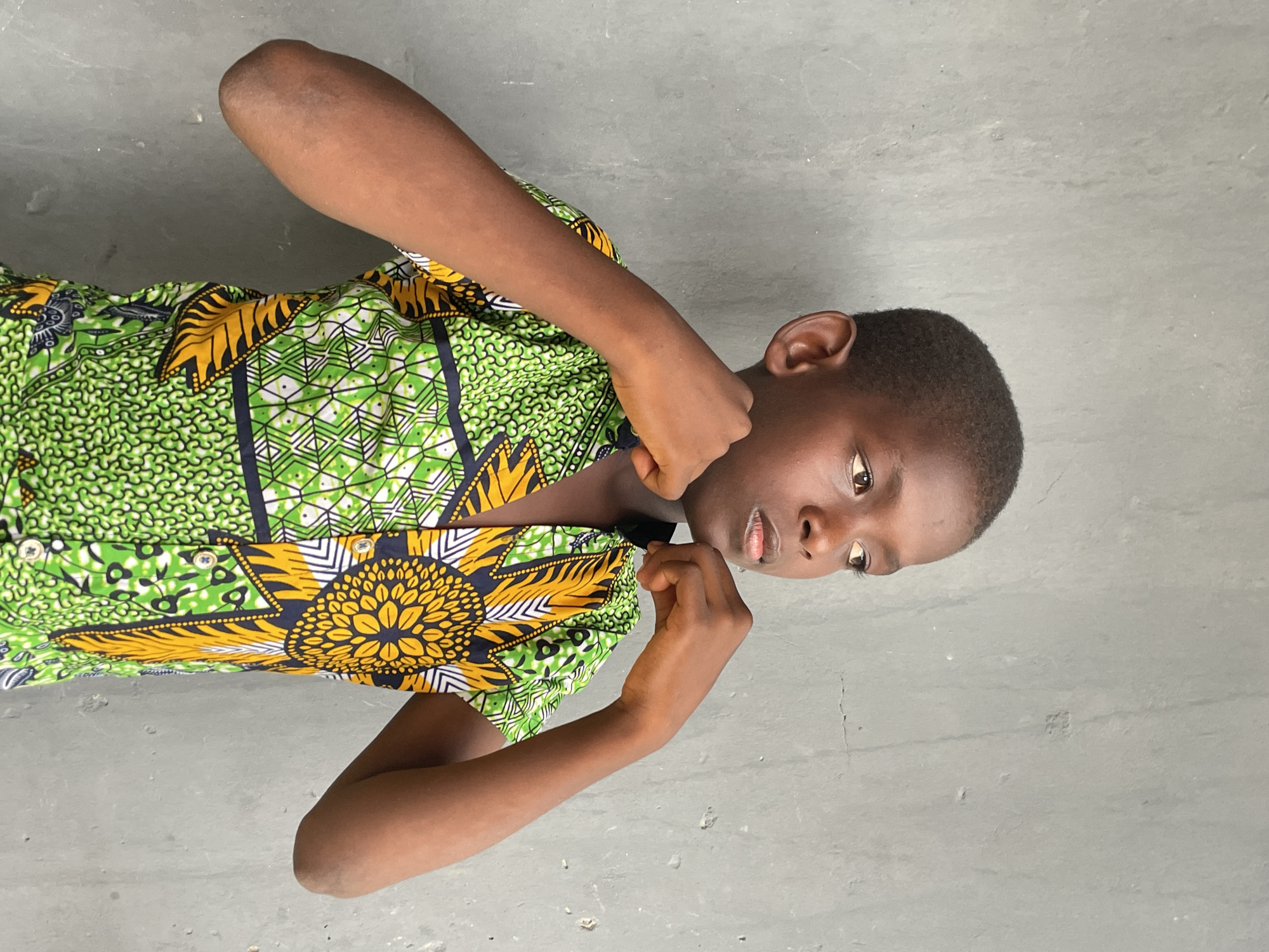 This portrait image, set in Badagry, Nigeria, features a young person, likely a boy, positioned against a plain, light grey background, which appears to be a concrete wall or floor. The subject has dark skin, short dark hair, and dark eyes, looking slightly to their right with a contemplative or somewhat serious expression.
They are wearing a vibrant, short-sleeved shirt with a bold pattern. The shirt is predominantly green, featuring intricate designs in white, black, yellow, and orange. A prominent motif on the shirt is a large, stylized yellow and black pattern resembling a sunflower or starburst, positioned near the chest and shoulder area. The subject's arms are bent at the elbows, with both hands clenched into fists and held up near their shoulders or chest, suggesting a pose of readiness, determination, or a playful stance. The lighting is bright and even, indicating it is daytime. There is no visible text in the image.
