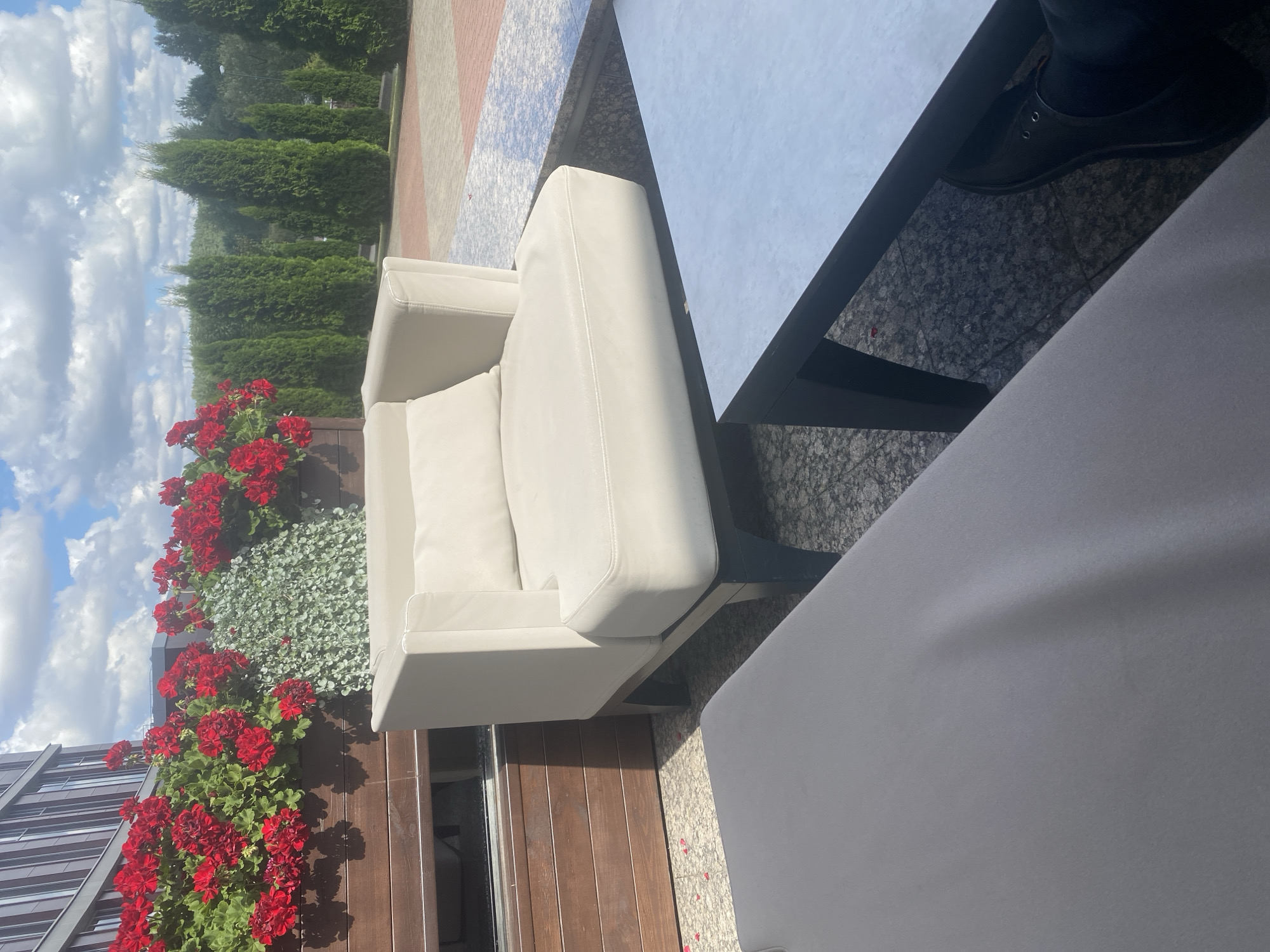 This image shows an outdoor seating area, likely a patio or terrace. In the foreground, a gray lounge chair or sofa is partially visible on the left. To the right of this, a light beige armchair with a single square pillow is prominently featured. Behind the armchair, a dark wooden planter box overflows with vibrant red geraniums and cascading green ivy. A modern building with a glass facade is visible in the upper left corner. Further back, a line of tall, slender green trees creates a backdrop. A rectangular coffee table with a light-colored top and dark legs sits in front of the armchair. The ground is paved with textured tiles. The sky above is blue with scattered white clouds. The scene appears to be set in Minsk, Belarus, as indicated by the specific architectural style and vegetation. There are no people visible in the image, but a pair of black dress shoes is seen peeking out from under the coffee table on the right side of the frame, suggesting someone is seated nearby. The overall impression is one of a tranquil outdoor space, possibly at a cafe, restaurant, or hotel.