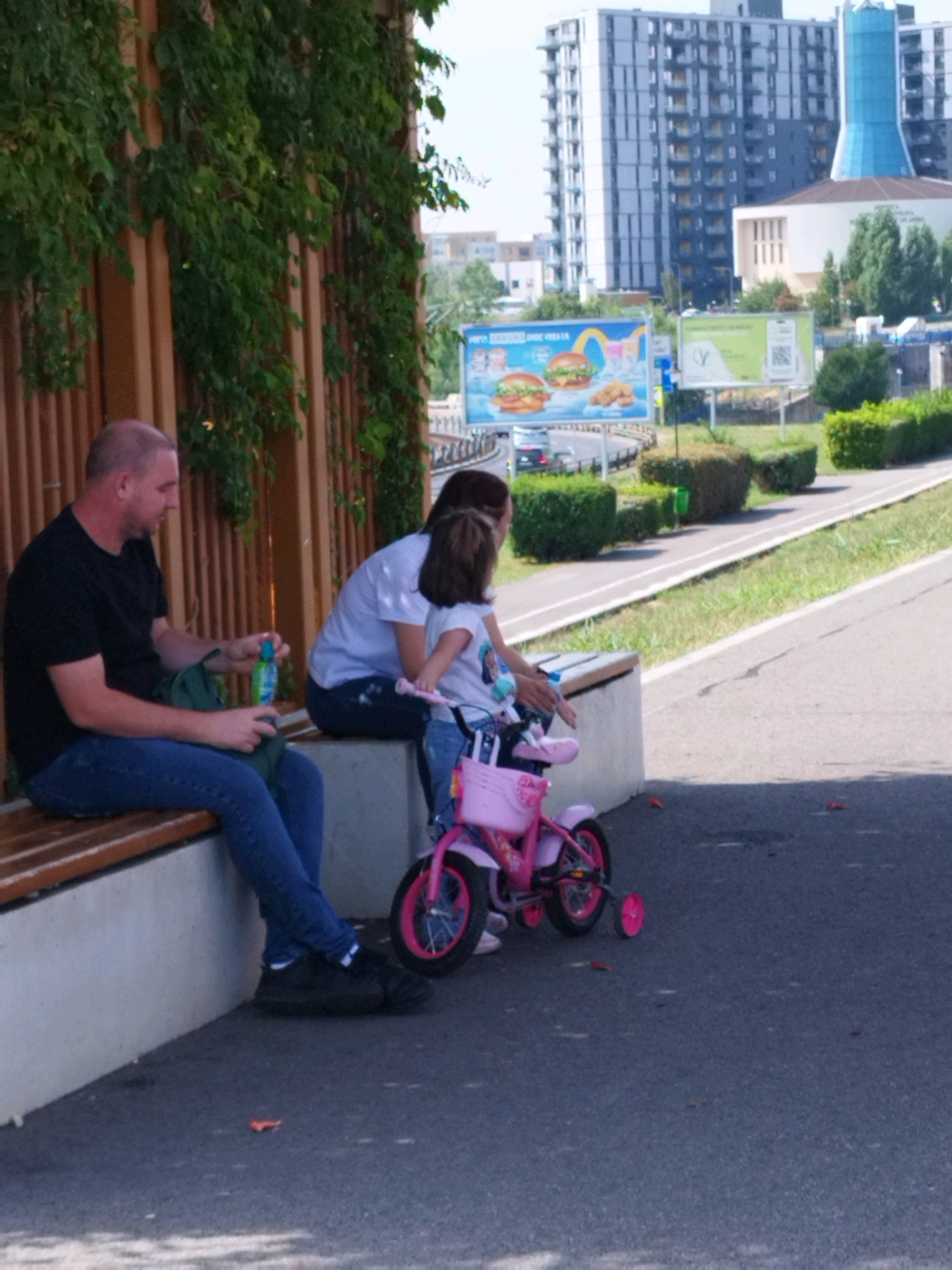 In Bucharest, Romania, a man and two children, a woman and a young girl, are seated on a bench near a pathway. The man, wearing a black t-shirt and blue jeans, is holding a green bag and a bottle of liquid. The woman, in a white t-shirt and blue jeans, is sitting next to him, and the young girl, in a white t-shirt with a graphic and blue jeans, is on her pink bicycle with training wheels. The background shows buildings, a billboard advertising food, and a bike path.