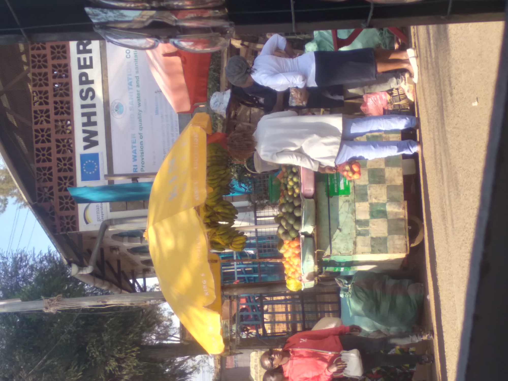 The image depicts an outdoor market scene, likely in Nyeri, Kenya, with several people present. A fruit stand is prominently featured, displaying a variety of fruits such as oranges and avocados under a yellow umbrella. Bananas hang from the stall.
To the right of the fruit stand, a woman in a red hoodie and dark pants is seen holding a white bag, appearing to be a customer. Further to the right, a man wearing a blue shirt and a patterned hat is interacting with another person. Two other individuals are visible near the stall, one in a white top and light blue pants, and another in a white long-sleeved shirt and a dark skirt.
In the background, a building with a sign is partially visible, indicating a project called "WHISPER" related to water and sanitation. The environment appears to be an urban or peri-urban setting with a paved road in the foreground. Bags of produce or other goods are stacked near the market stall. The overall scene suggests a typical day at a local market.