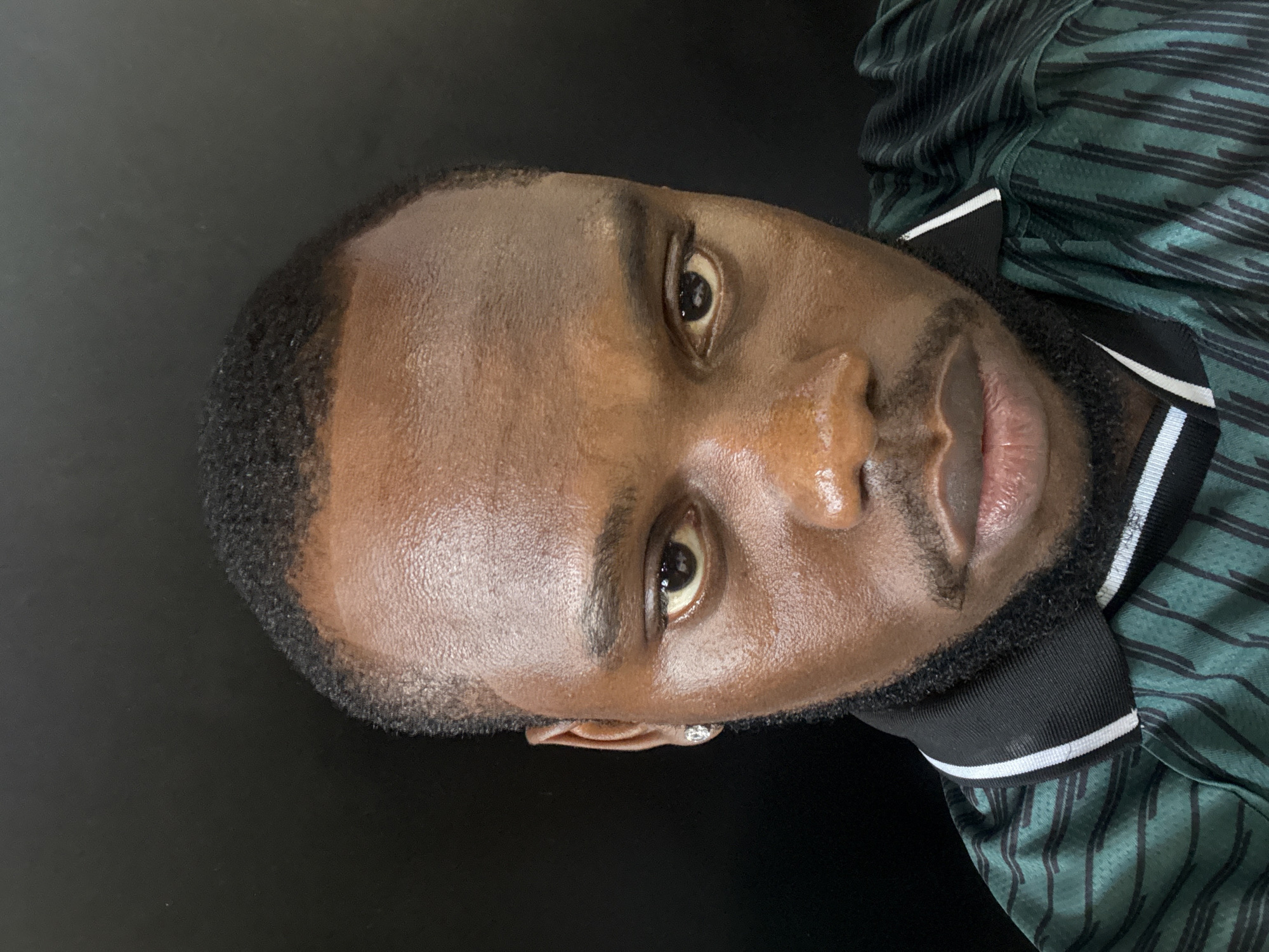 A headshot of a Black man. He has dark eyes, a short haircut, and a beard. He is wearing a green shirt with black stripes and a black and white collar. The background is a plain black surface. There is no evidence to indicate the city or location of Lugbe, Nigeria.