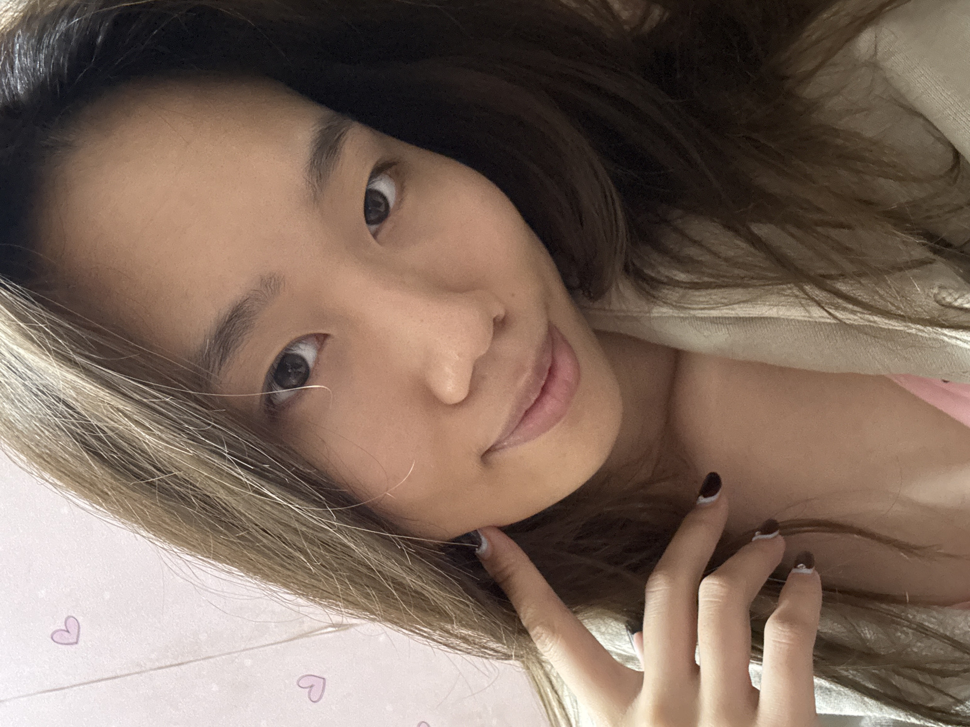 A selfie of a light-skinned Asian woman with long, ombre brown hair. She is tilted towards the right with her head in her hand with brown and white nail polish on her nails. The background is a light pink color with small heart shapes. The location is unknown and there is no way to determine if this picture was taken in Shapingba District, China or anywhere else.