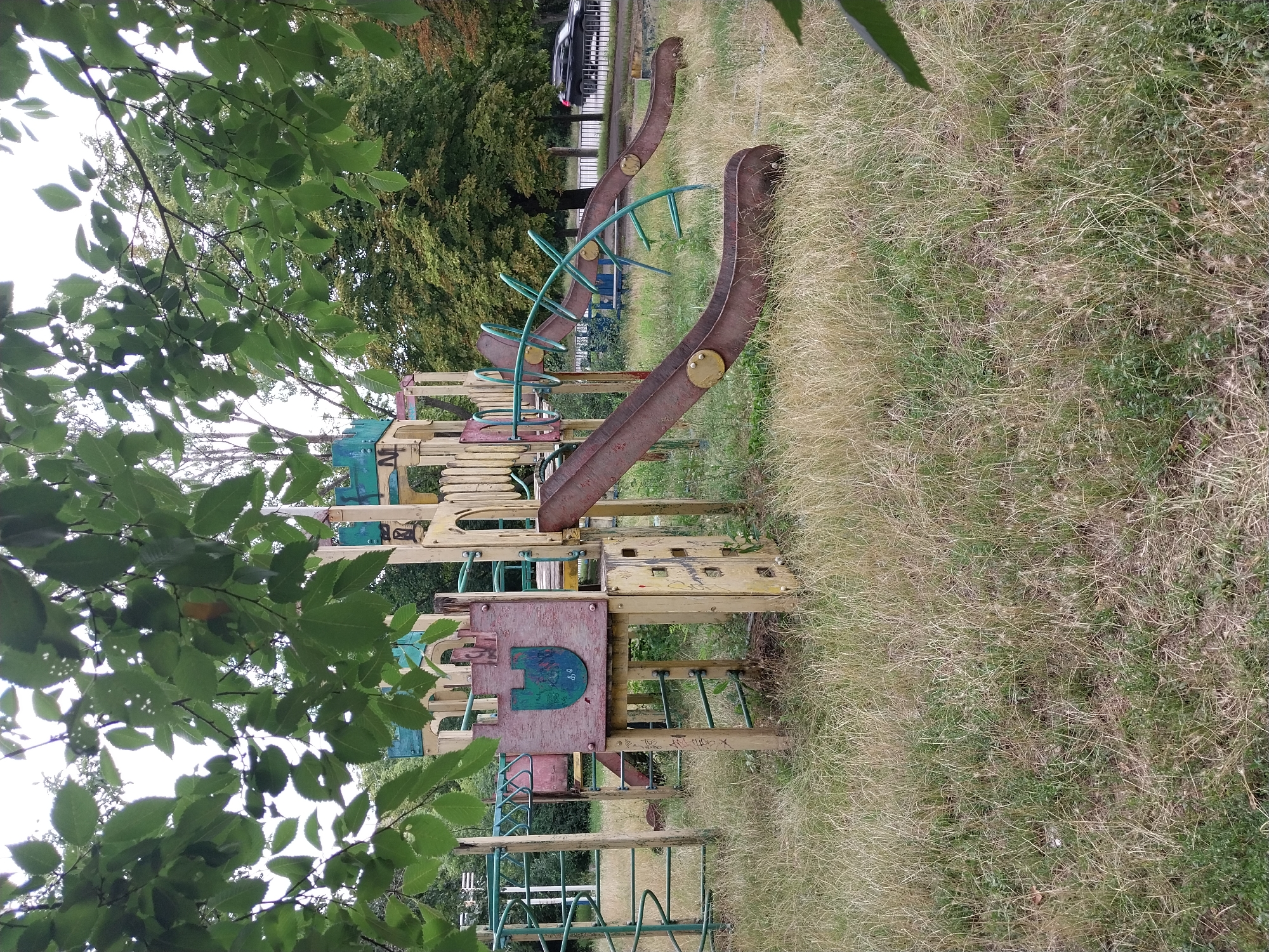 An abandoned, dilapidated children's playground is situated in an overgrown grassy area. The playground structure, made of wood and metal, appears weathered and rusted. It features slides, climbing structures, and a central platform decorated with a shield emblem. The surrounding vegetation is unkempt, with dry grass and overhanging tree branches obscuring parts of the scene. In the background, a road with a passing car and a metal fence can be seen, suggesting an urban setting. The overall impression is one of neglect and disuse. This scene is located in the city of Dnipro, Ukraine.