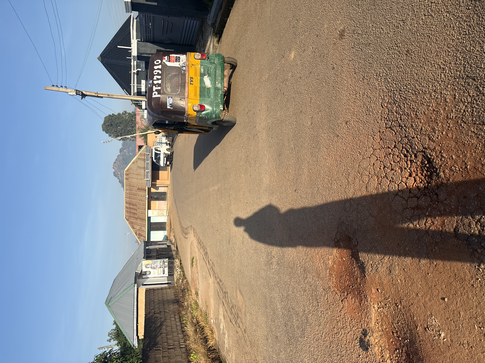 In Bukuru, Nigeria, on a clear, sunny morning, a shadow of a person stands prominently on a paved road, stretching long across the cracked asphalt. The road curves gently to the right, leading towards buildings on both sides. On the left, a brick wall runs alongside the road, bordering a property with a metallic, sloped roof. Further down the road, a building with a similar roof design is visible.
On the right side of the road, a three-wheeled vehicle, commonly known as a Keke Napep, is parked, its rear facing the camera. The vehicle is green and yellow and displays the license plate "PT-17910". A utility pole with wires extends upwards behind the vehicle. Beyond the Keke Napep, a house with a distinctive black, conical roof is visible, followed by a grey metal gate.
In the distance, a white truck is seen moving away from the camera, and beyond that, a rocky hillside under a clear blue sky suggests the natural landscape surrounding the town. The overall scene captures a typical urban setting in Bukuru, with elements of daily life and the characteristic architecture of the region under the morning sun.