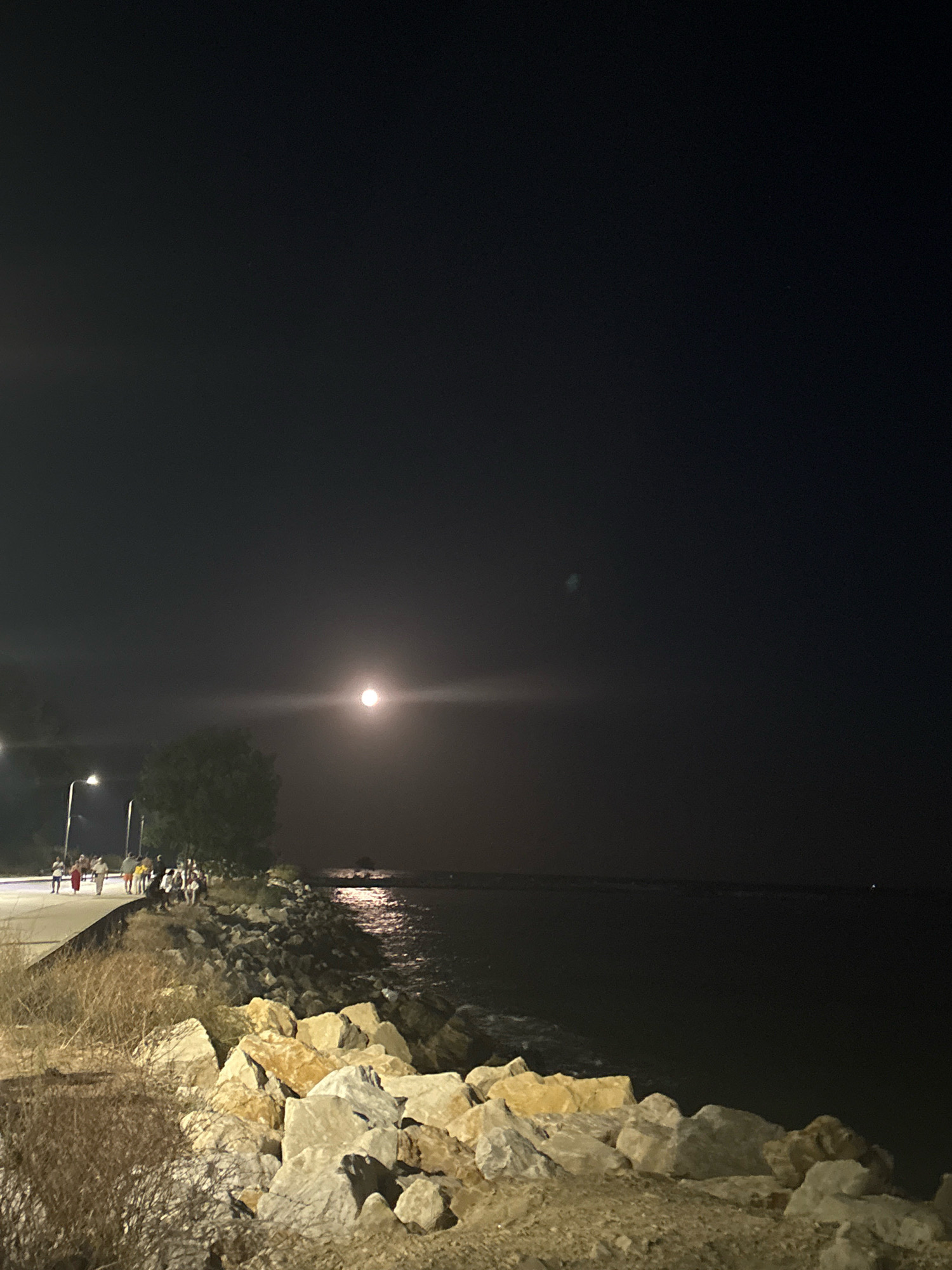 A nighttime, medium-shot photograph captures a waterfront scene under a full moon. The moon, a bright, luminous orb, hangs in the dark, hazy sky, casting a faint glow and a bright horizontal streak across the upper portion of the frame. In the foreground, a rocky embankment made of large, light-colored stones lines the edge of the dark water. Dry grass and brush grow on the left side of the embankment.
A paved walkway runs alongside the embankment, populated by a group of approximately ten to fifteen people walking and standing. Streetlights illuminate the walkway, casting pools of light. The water on the right appears dark and calm, with a subtle reflection of distant lights shimmering on its surface. The overall atmosphere is serene, with the moonlit night creating a slightly diffused and ethereal quality to the scene. The exact location in the city of Unknown, Unknown is not discernable from the image.