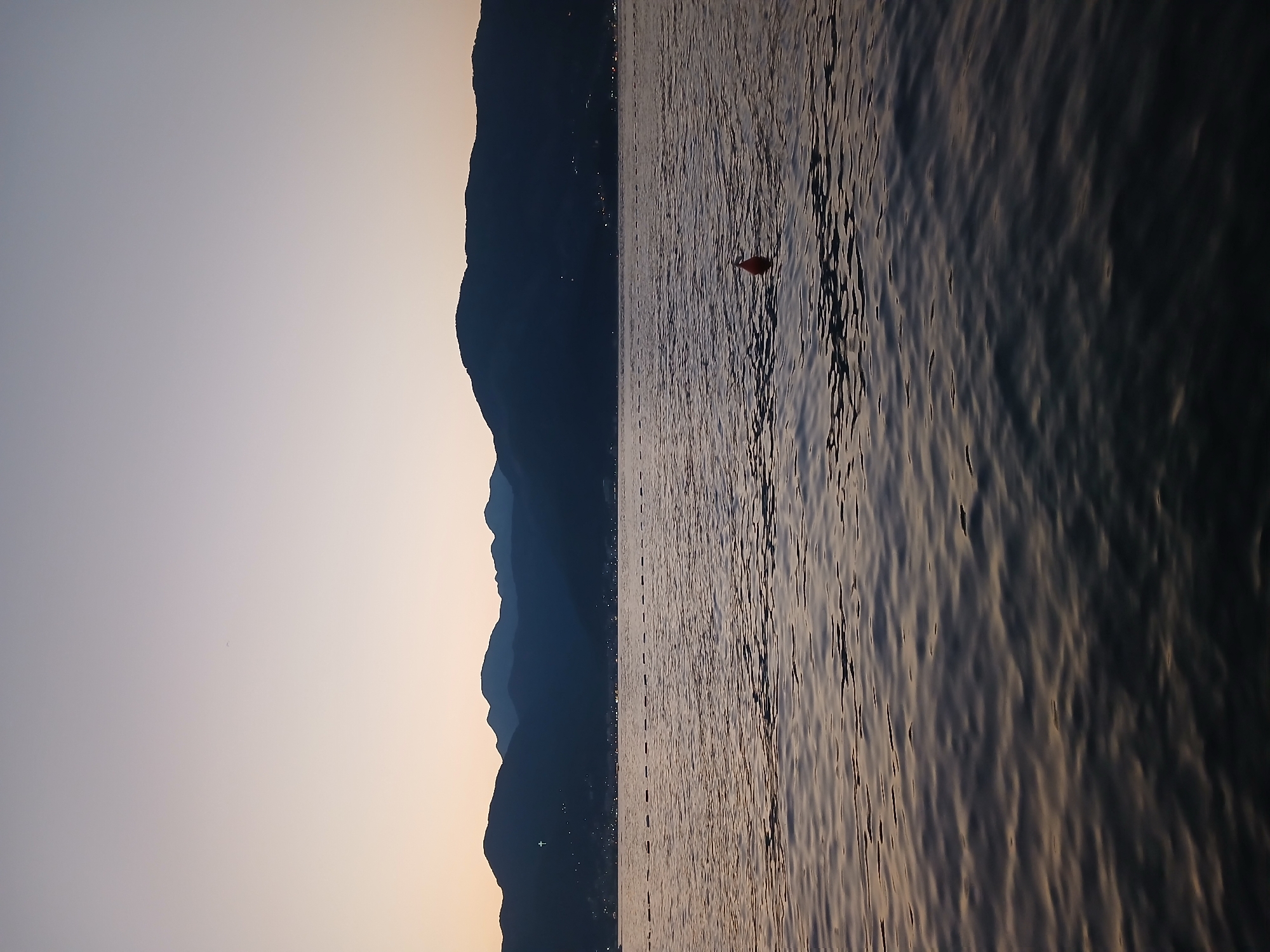 The image depicts a body of water with a mountainous coastline in the background. The sky is a gradient of soft colors, transitioning from pale yellow at the horizon to a muted gray at the top of the frame, suggesting either sunrise or sunset. Gentle waves are visible on the water's surface, reflecting the soft light. A single, small orange buoy floats in the water towards the right side of the frame. In the distance, along the base of the mountains, a faint line of lights suggests a settlement or town. The mountains themselves are dark silhouettes against the lighter sky, their forms layered and indistinct. No people or specific landmarks are clearly discernible, giving the scene a tranquil and somewhat remote feel. The overall impression is one of a peaceful natural landscape at the cusp of day or night.