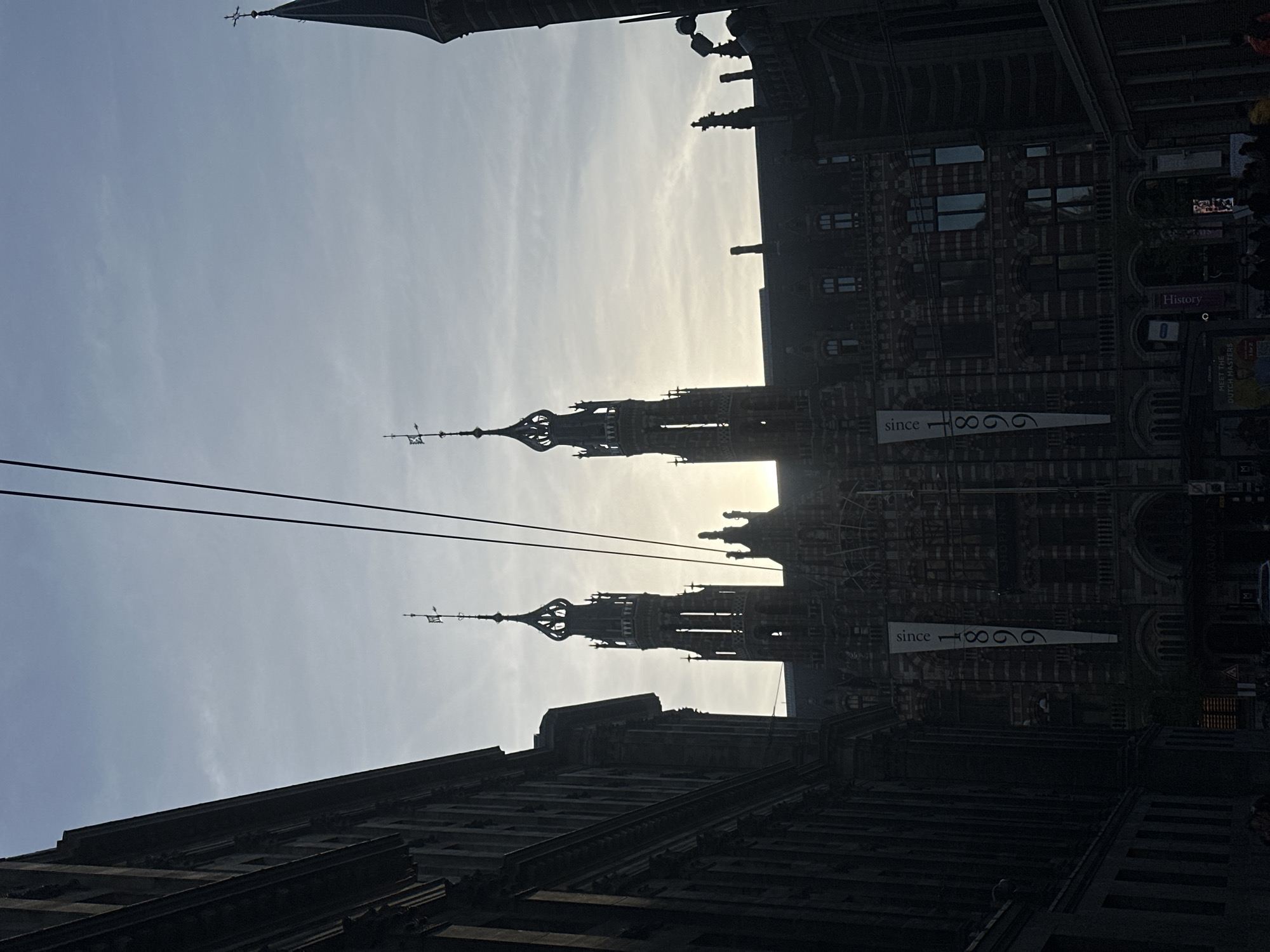 The image shows a low-angle view of the Beurs van Berlage in Amsterdam, Netherlands. The architecture is prominently featured, with its characteristic towers and intricate detailing silhouetted against a bright sky. A banner with the inscription "since 1866" is visible on the building. Power lines cut across the sky in the foreground. People are present on the right side of the frame, with a sign indicating "History". The overall environment seems to be a bustling urban area, captured in a vertical orientation.