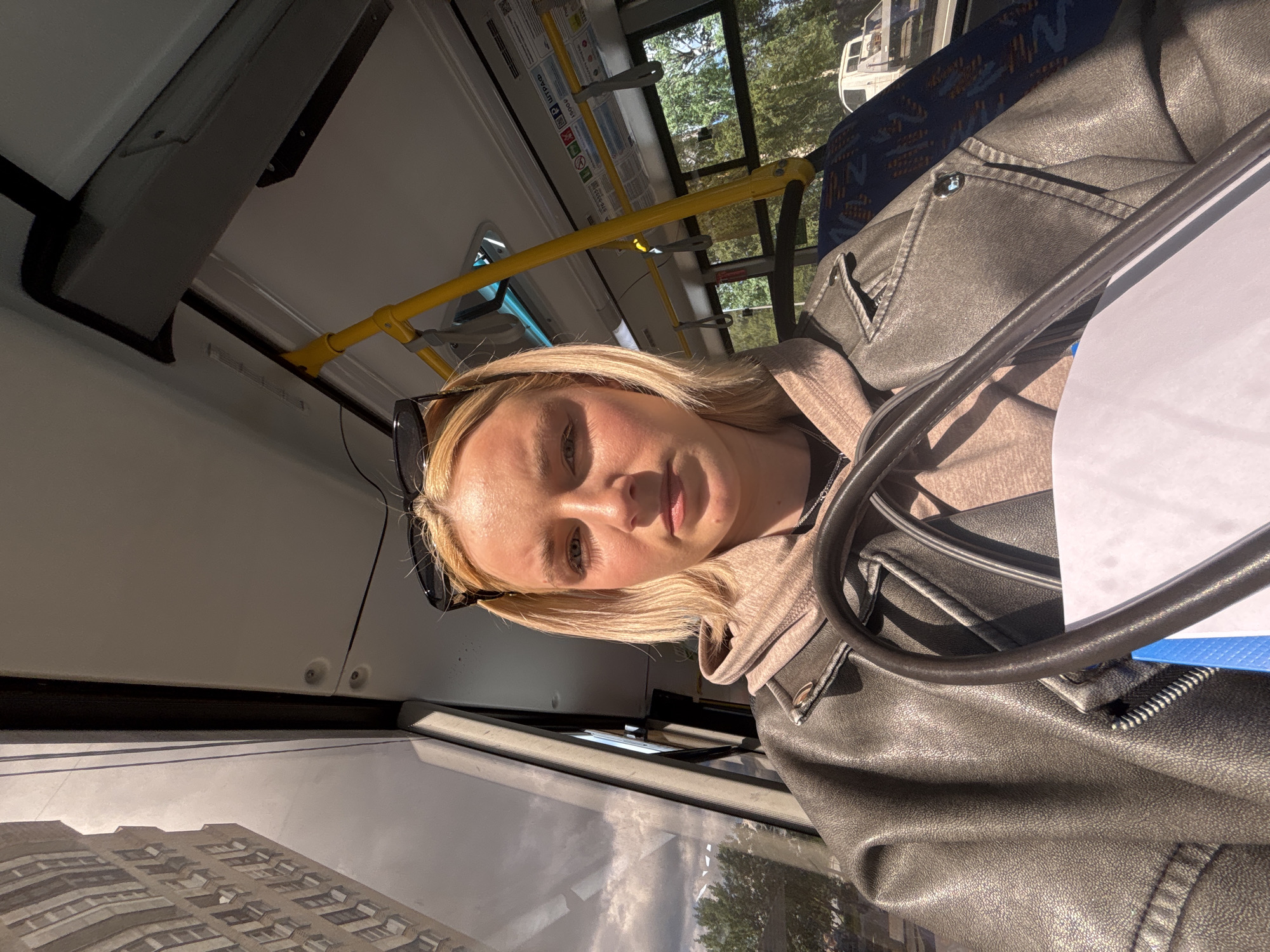 The media file is a selfie taken by a young woman with blonde hair inside what appears to be a public transportation vehicle, possibly a bus or tram. She is wearing a light brown hooded sweatshirt under a dark gray leather jacket. Sunglasses are perched on her head. The setting is illuminated by natural light, suggesting it is daytime, likely with clear skies given the bright sunlight casting shadows and highlighting the textures of her clothing.
Through the windows, trees and a white vehicle are visible, indicating an outdoor environment. Reflections on the window show a building, potentially an apartment complex, suggesting an urban or suburban location. The interior of the vehicle shows blue and gray patterned seating and yellow grab handles, common features in public transport.
The woman's facial expression is neutral, with her gaze directed towards the camera. Her skin has a warm tone, possibly from the sunlight. The image is oriented sideways, suggesting it was taken with a mobile phone held vertically. No explicit text is clearly visible or legible in the image. The overall mood is casual and everyday, capturing a moment during a commute.