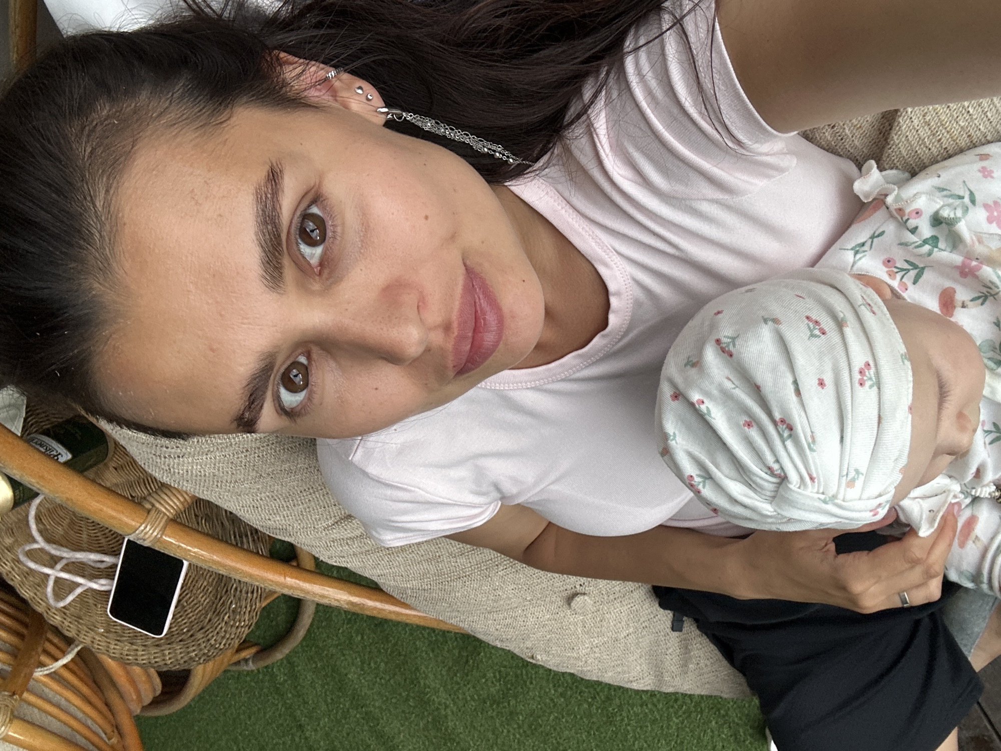 A woman with dark hair and brown eyes is holding a baby. The woman is wearing a pink t-shirt and black pants. The baby is wearing a white hat with a floral pattern and a white onesie with a floral pattern. They are sitting on a chair covered with a beige textured fabric. To the left of the chair, there is a woven basket with a white cord and a smartphone on it. The background appears to be an outdoor setting with green artificial turf. The scene is illuminated by natural light. The location is in the city of Sylva, Russia.