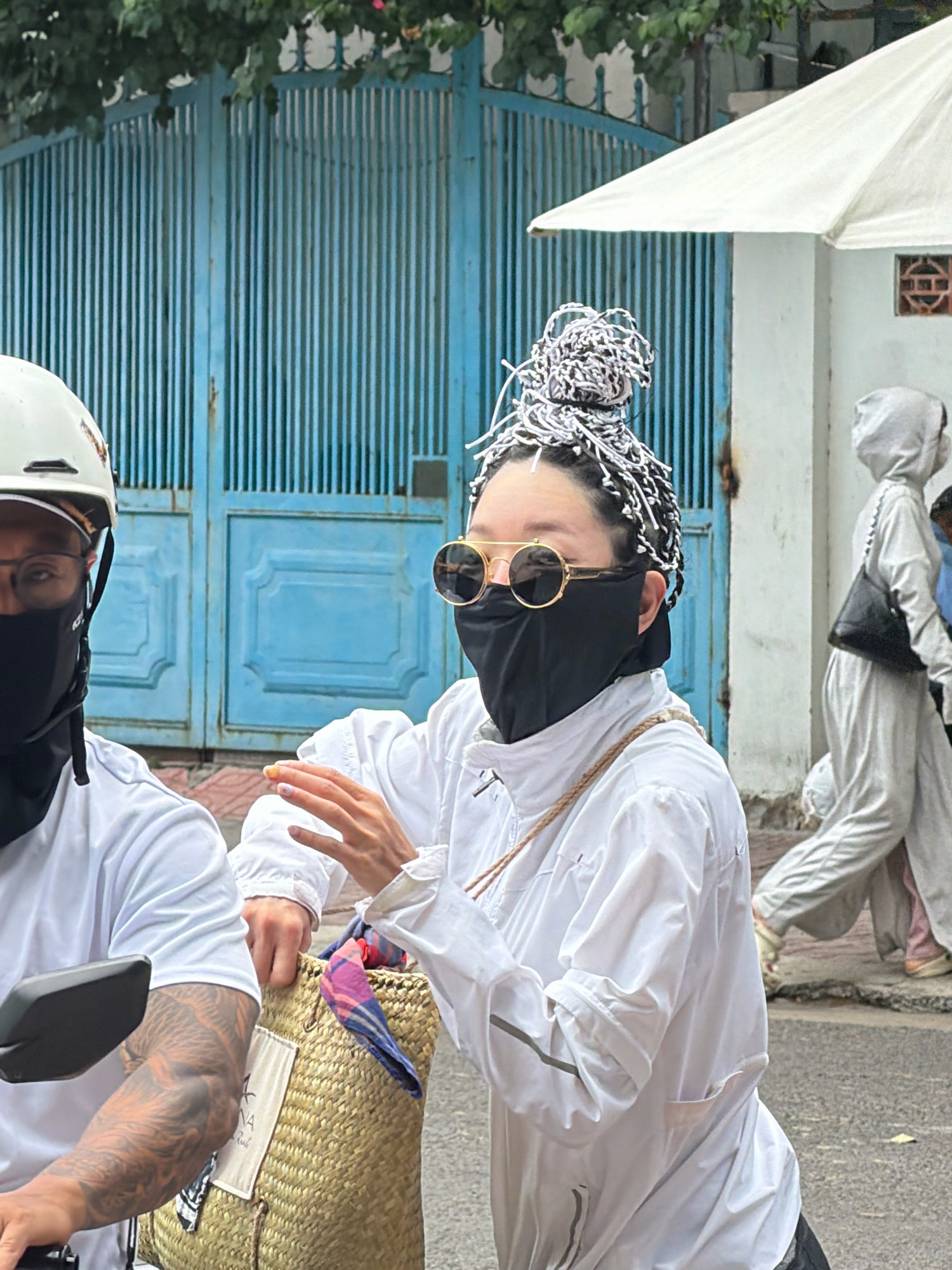 In Nha Trang, Vietnam, a woman with a distinctive braided hairstyle is captured from the chest up, wearing a black face mask, round sunglasses with gold frames, and a white hooded jacket. Her left hand is raised, with fingernails painted in a colorful, abstract design. She is holding a woven straw bag with a colorful scarf peeking out. To her left, partially visible, is a person wearing a white helmet and a black face mask, with a white t-shirt and tattoos visible on their arm. In the background, a blue gate with vertical slats is prominent, with green foliage above it. To the right, another person in a light-colored hooded outfit with a black purse is walking away. A white canopy is visible overhead. The scene appears to be on a street.