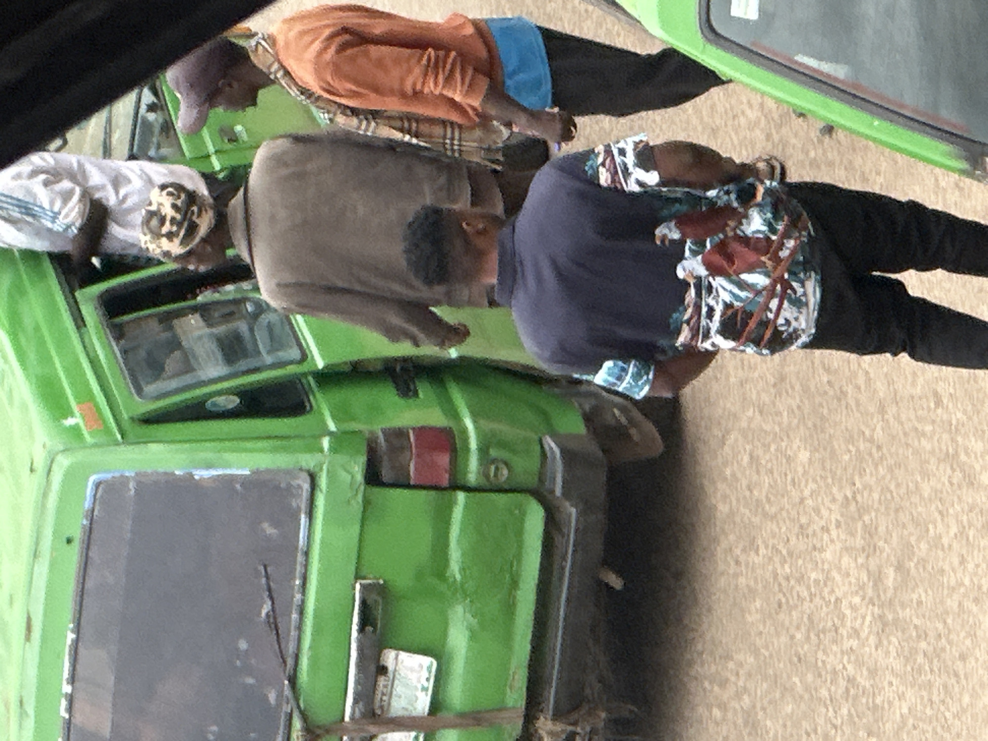 This image depicts a street scene in Bukuru, Nigeria, focusing on the side and rear of a bright green van. Several individuals are present, interacting with the vehicle. In the foreground, a man wearing a dark blue polo shirt with a colorful eagle print is facing away from the camera, his back to the van. To the right of him, another man in an orange long-sleeved shirt and a purple baseball cap is also facing away, looking towards the van. Behind the first man, a person in a brown jacket and a patterned headscarf is standing near the open door of the van. Another individual, mostly out of frame, is wearing a white shirt with blue stripes and appears to be entering or exiting the van. The van itself is a prominent green color, showing some signs of wear and tear. The ground appears to be a dusty, unpaved surface, typical of a street or informal parking area. The overall impression is one of everyday activity, possibly related to transport or commerce, on a street in Bukuru.