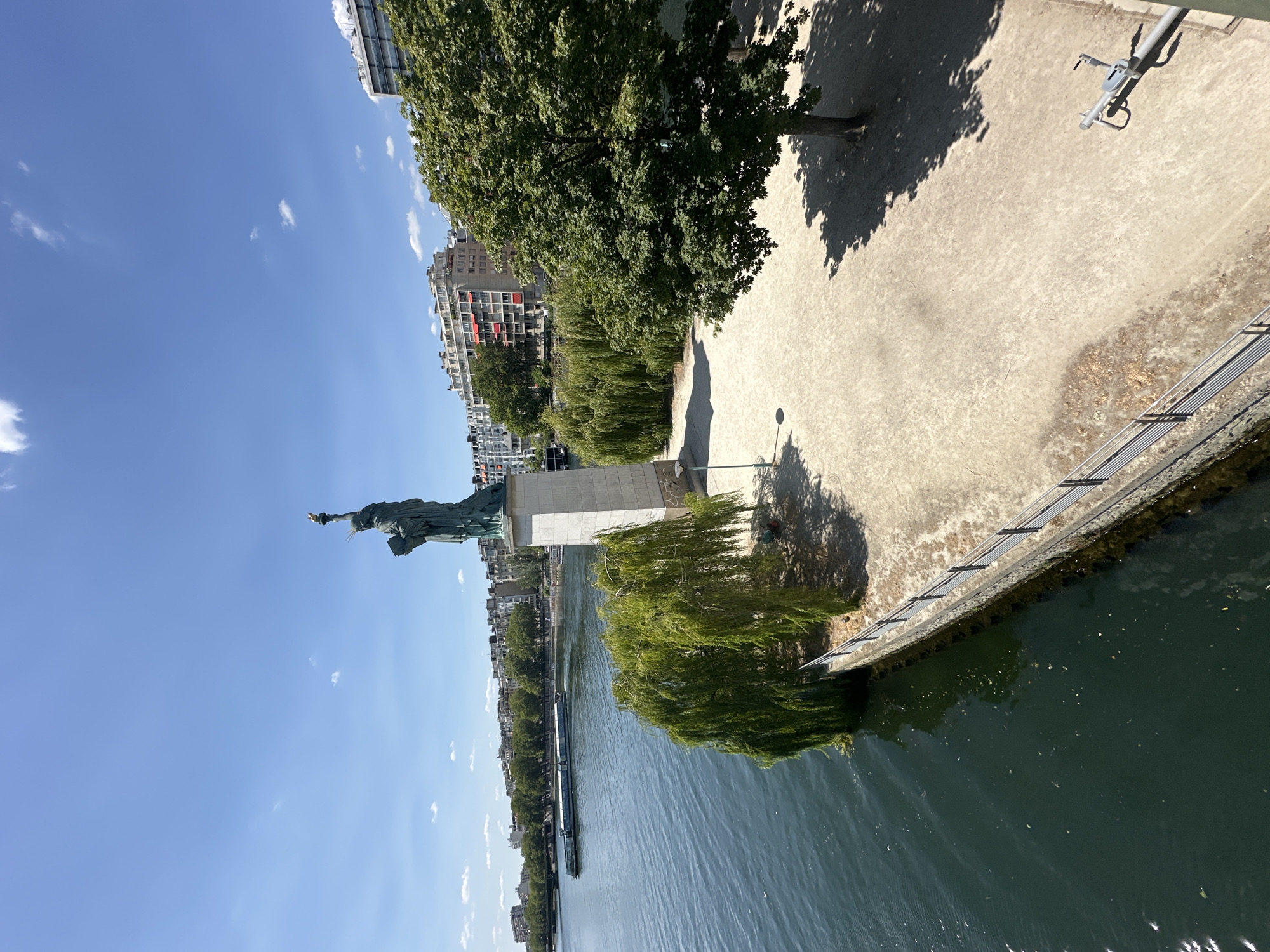The image shows a view of the Statue of Liberty replica on the Île aux Cygnes in Paris, France. The statue stands on a white pedestal on the left side of the image. A body of water, likely the Seine River, is in the foreground, with a tour boat navigating on it. On the right side of the image, a paved area with trees and shadows is visible. In the background, apartment buildings and greenery line the opposite bank of the river. The sky is clear and blue with a few scattered clouds. A bicycle rack is visible on the paved area in the lower right corner.
