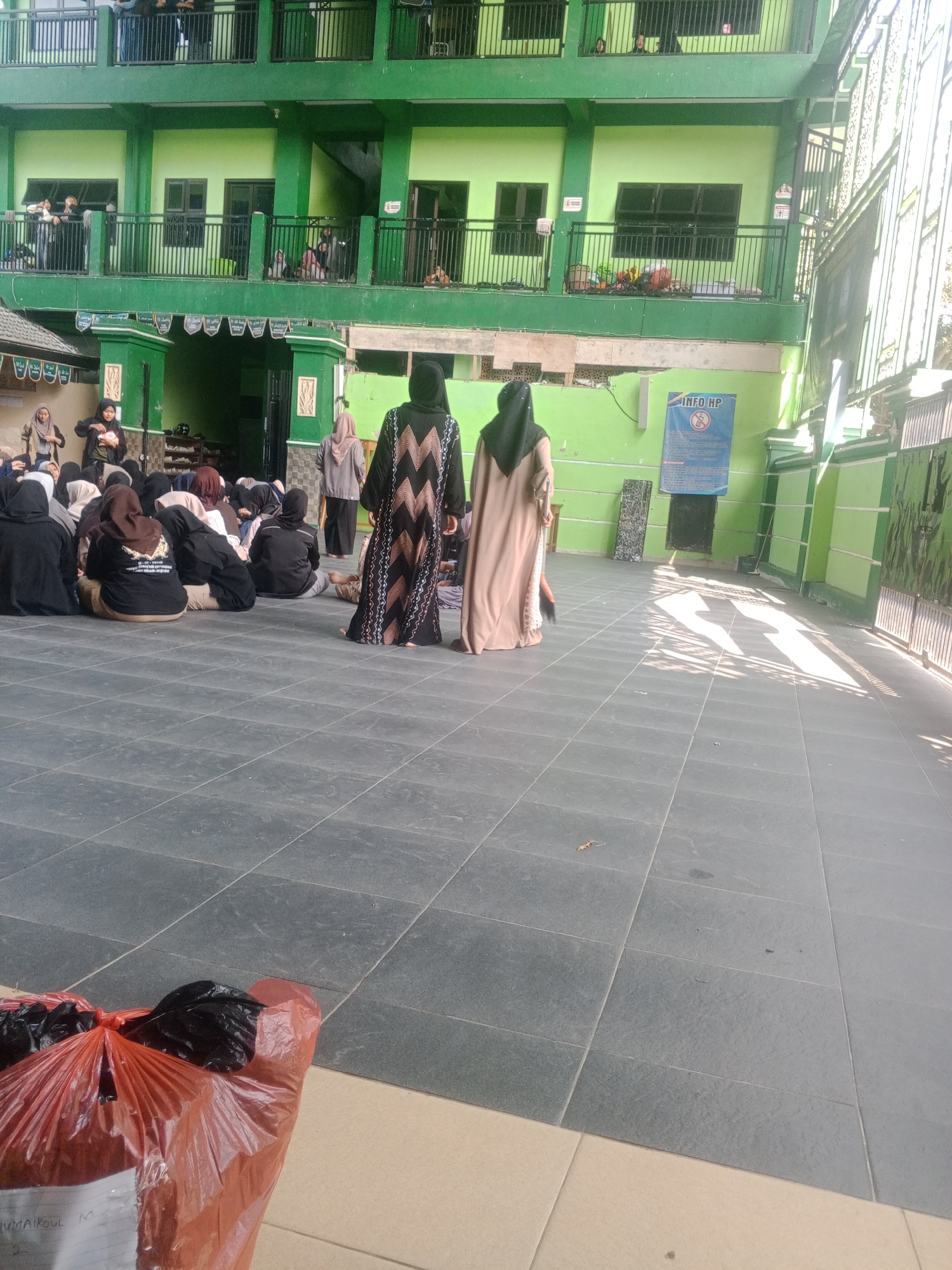 In Banyudono, Indonesia, a group of individuals, predominantly women wearing hijabs and Islamic attire, are gathered outdoors on a tiled courtyard. They are seated on the ground, facing the entrance of a multi-story green building, which appears to be a school or religious institution. The building has multiple levels with balconies and windows, and some people can be seen on the balconies. One woman in a patterned black and white abaya and another in a light brown abaya are standing in the foreground, facing away from the camera. To the left, a large orange plastic bag filled with dark items is visible on the ground. The courtyard is paved with dark gray tiles, and sunlight casts distinct shadows across the area. A sign with "INFO HP" is posted on the building's exterior wall. The overall scene suggests a gathering or event taking place in an educational or community setting.