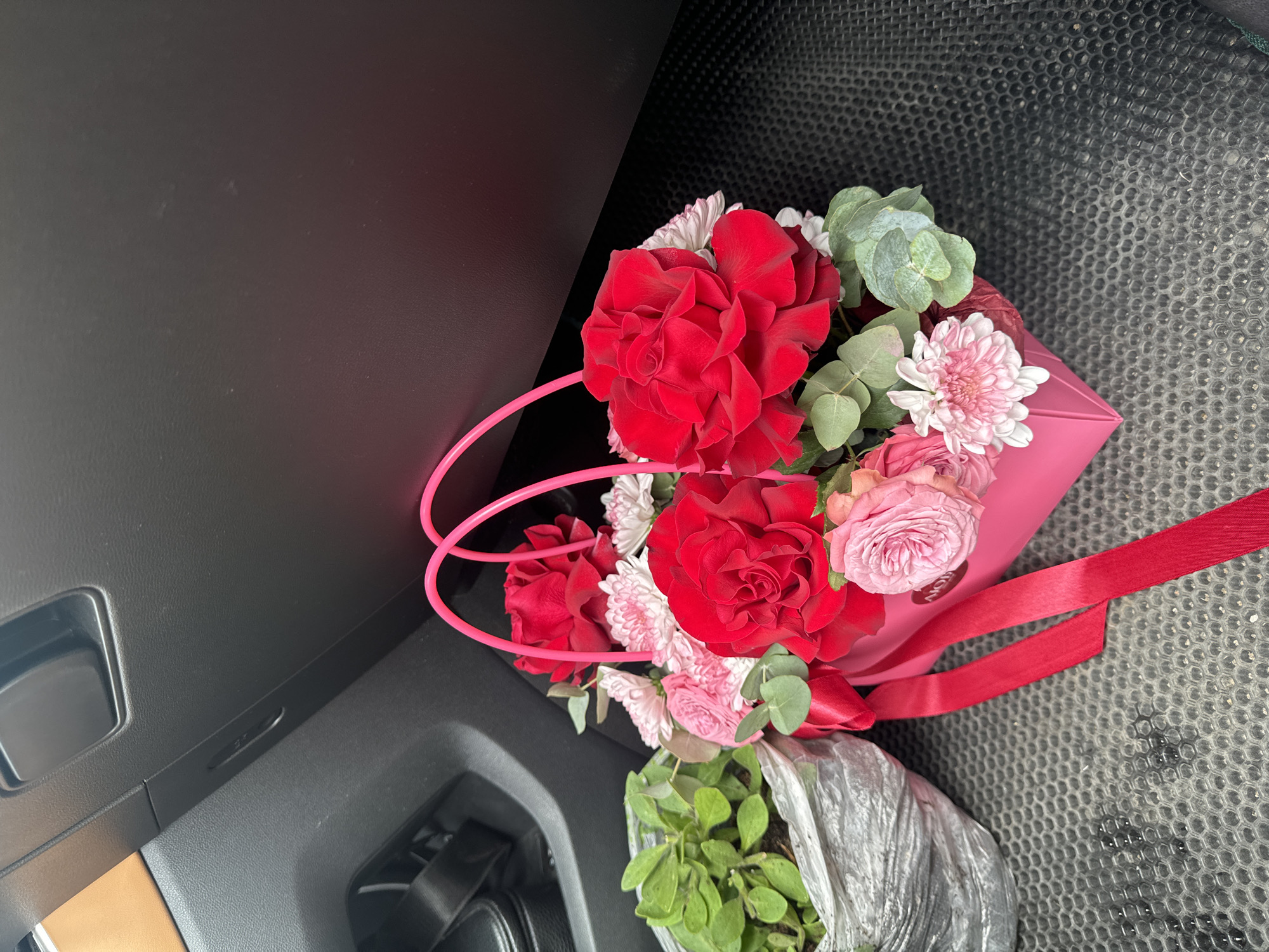 A close-up, eye-level shot captures a bright pink flower arrangement, featuring vibrant red roses and delicate pink roses, nestled within a pink box with matching handles. The arrangement is adorned with a bright red ribbon. To the left of the flower arrangement, a small potted plant in a textured, gray wrapper is visible. The scene appears to be inside a vehicle, with the black dashboard and textured floor mat clearly visible. The background suggests the interior of a car, with a portion of a tan dashboard and a black seat visible on the left. The lighting is moderate, highlighting the colors of the flowers. The image does not contain any information about the location in the city of Сароцкі сельскі Савет, Belarus.