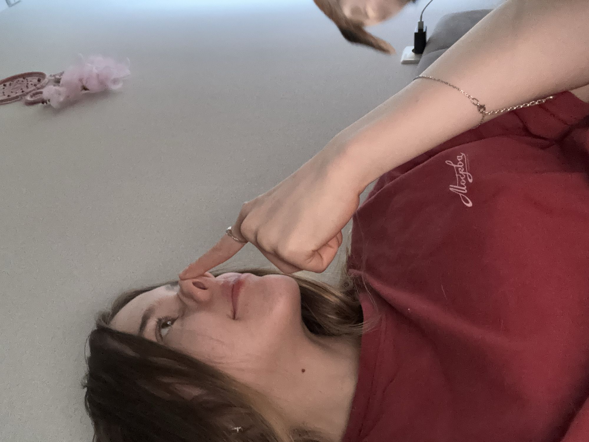 The image shows a close-up of a young woman's face and upper body. She has medium-brown hair, and she is looking up and to her right. Her right index finger is touching her nose, and she is wearing a ring on that finger. She is wearing a dark red t-shirt with a white embroidered logo that appears to say "Москва" (Moscow in Russian). A delicate bracelet is visible on her left wrist. In the background, a pink fluffy decoration hangs on a light-colored wall. To the right of the woman, out of focus, is a brown dog's ear and a glimpse of its face, and a white electrical outlet with a black plug. The scene suggests an indoor domestic setting.