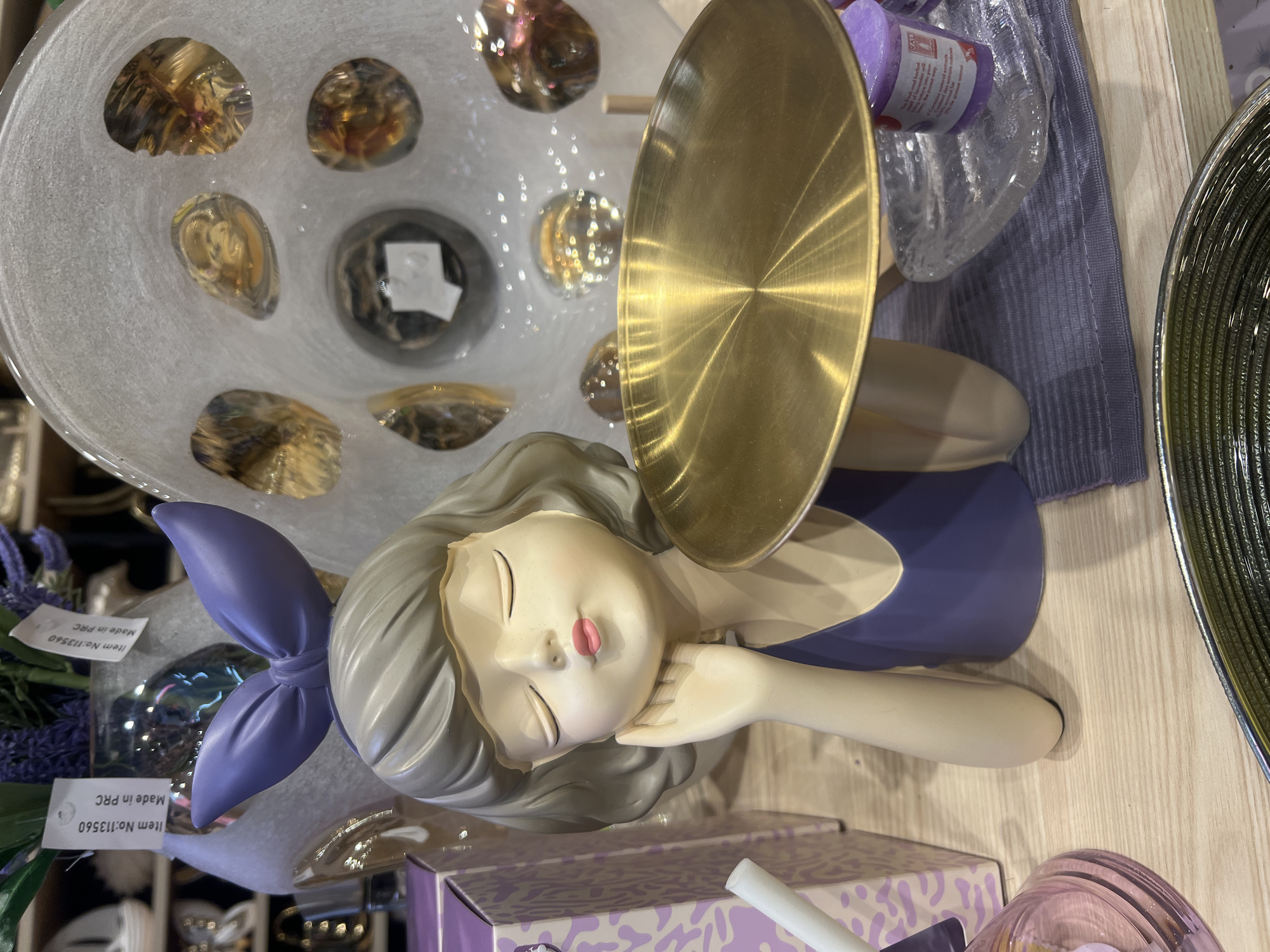 A figurine of a young girl with closed eyes and a purple bow on her head rests her cheek on her hand. She wears a purple dress and holds a gold tray. She is placed on a wooden surface, with a purple box and a decorative glass vase with amber-colored stones in the background. The setting appears to be a retail environment, potentially a gift shop or home decor store. The location is specified as Lisky, Ukraine.