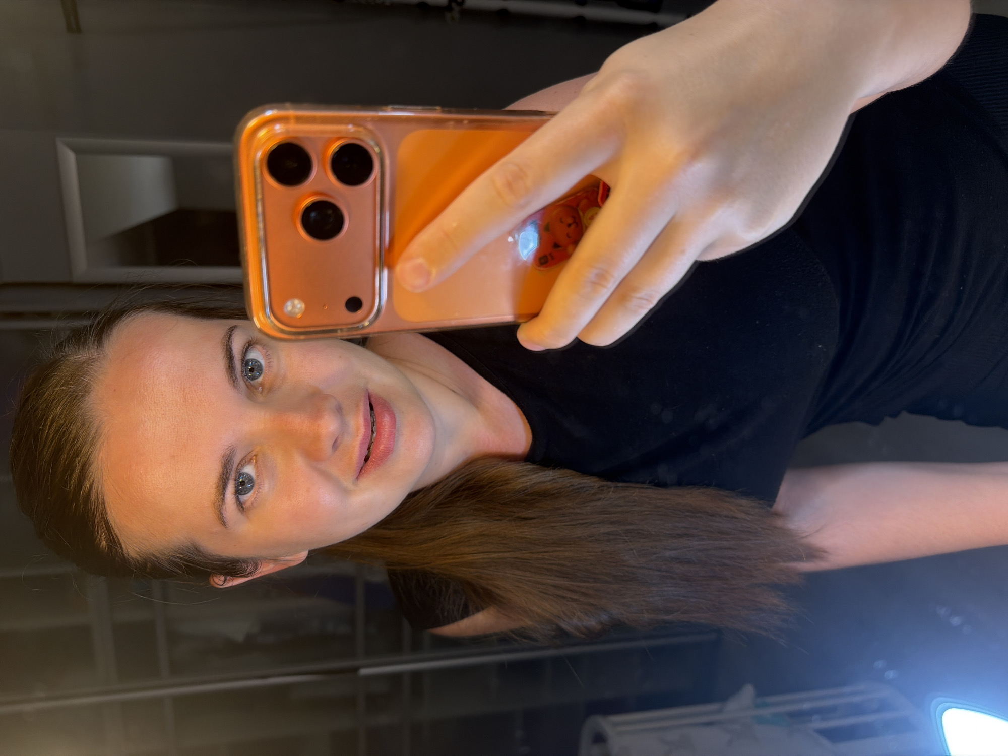 The image displays a selfie of a young woman, taken indoors in Sochi, Russia. The woman has fair skin, clear blue eyes, and long, straight brown hair pulled back into a ponytail that falls over her right shoulder. She looks directly at the camera with a gentle, engaging smile. She is wearing a simple black top.
She holds a smartphone in her right hand, prominently positioned in the upper right quadrant of the frame. The phone, identifiable by its triple camera array, is housed in a translucent, orange-tinted protective case. A subtle reddish-orange character or design is visible on the back of the phone case, underneath her fingers.
The background is dimly lit and somewhat obscured, featuring architectural elements such as recessed panels or shelves, indicating an indoor environment, possibly a room or a storage area. A strong light source emanates from the lower right corner, creating a bright glare that reflects off a surface. The photograph is oriented vertically but is displayed rotated 90 degrees counter-clockwise. No legible text is visible within the image. The indoor setting and artificial lighting prevent determination of the time of day or outdoor weather conditions.