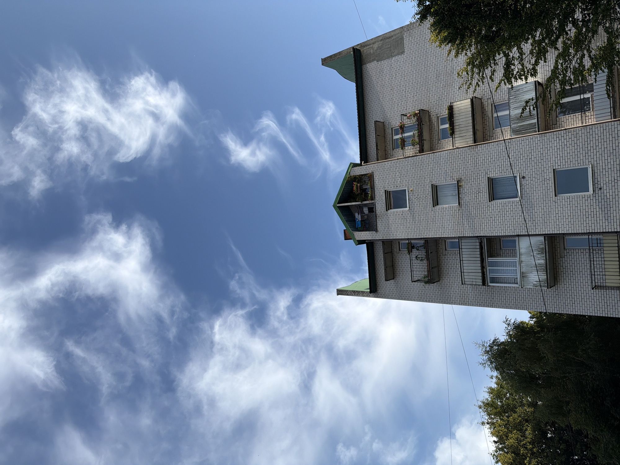 A low-angle, exterior shot captures the upper floors of a white brick apartment building against a blue sky with wispy white clouds. The building has a green, pitched roof and multiple balconies with metal railings, some of which have potted plants. Trees are visible to the left of the building, and power lines are strung across the frame. The scene appears to be in Kaliningrad, Russia. There are no people visible in the image.