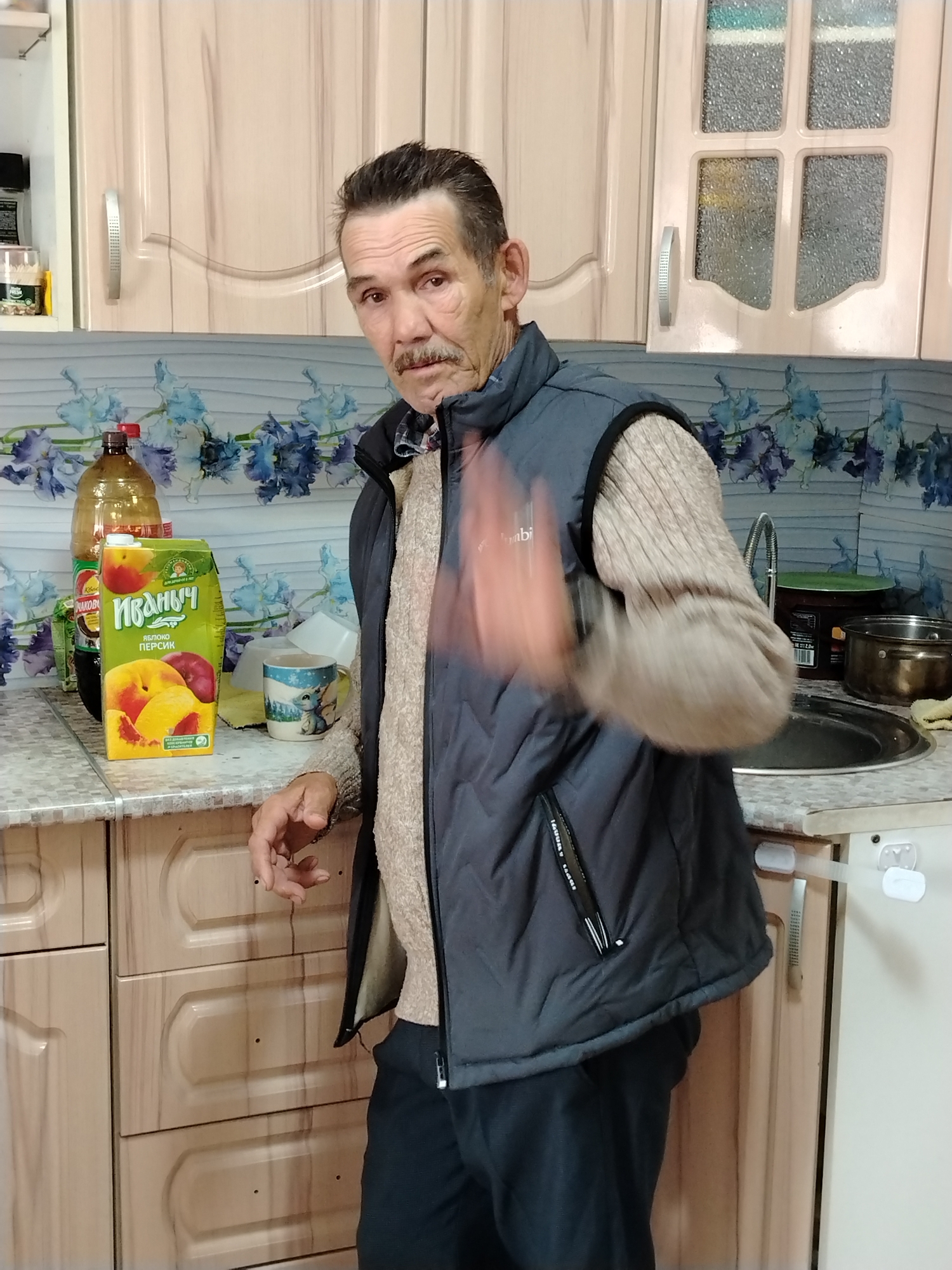 An older man with a mustache, wearing a sweater and a dark vest, stands in a kitchen and waves his hand. The kitchen features light-colored cabinets with decorative paneling, a countertop with various items including a carton of juice, a bottle, and a mug, as well as a sink and pots. The backsplash is decorated with a floral pattern. The location is Unknown, Unknown.