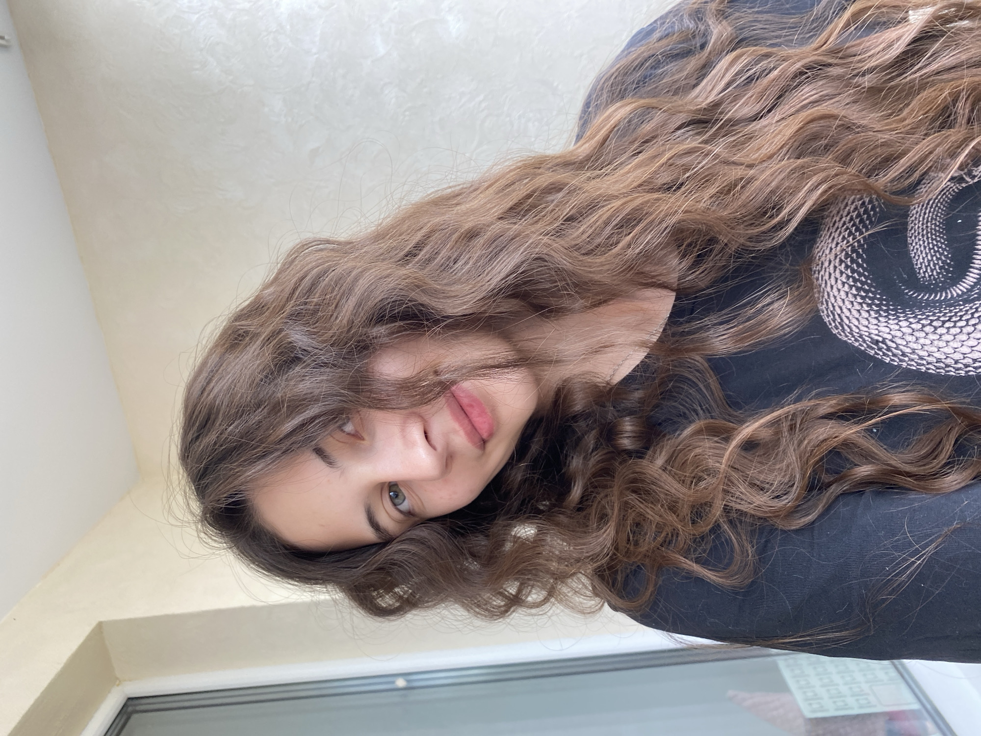A young woman with long, wavy brown hair is pictured. She is wearing a black t-shirt with a snake design. The background consists of a light-colored wall and what appears to be a window. There is no information to suggest the image was taken in Votkinsk, Russia.