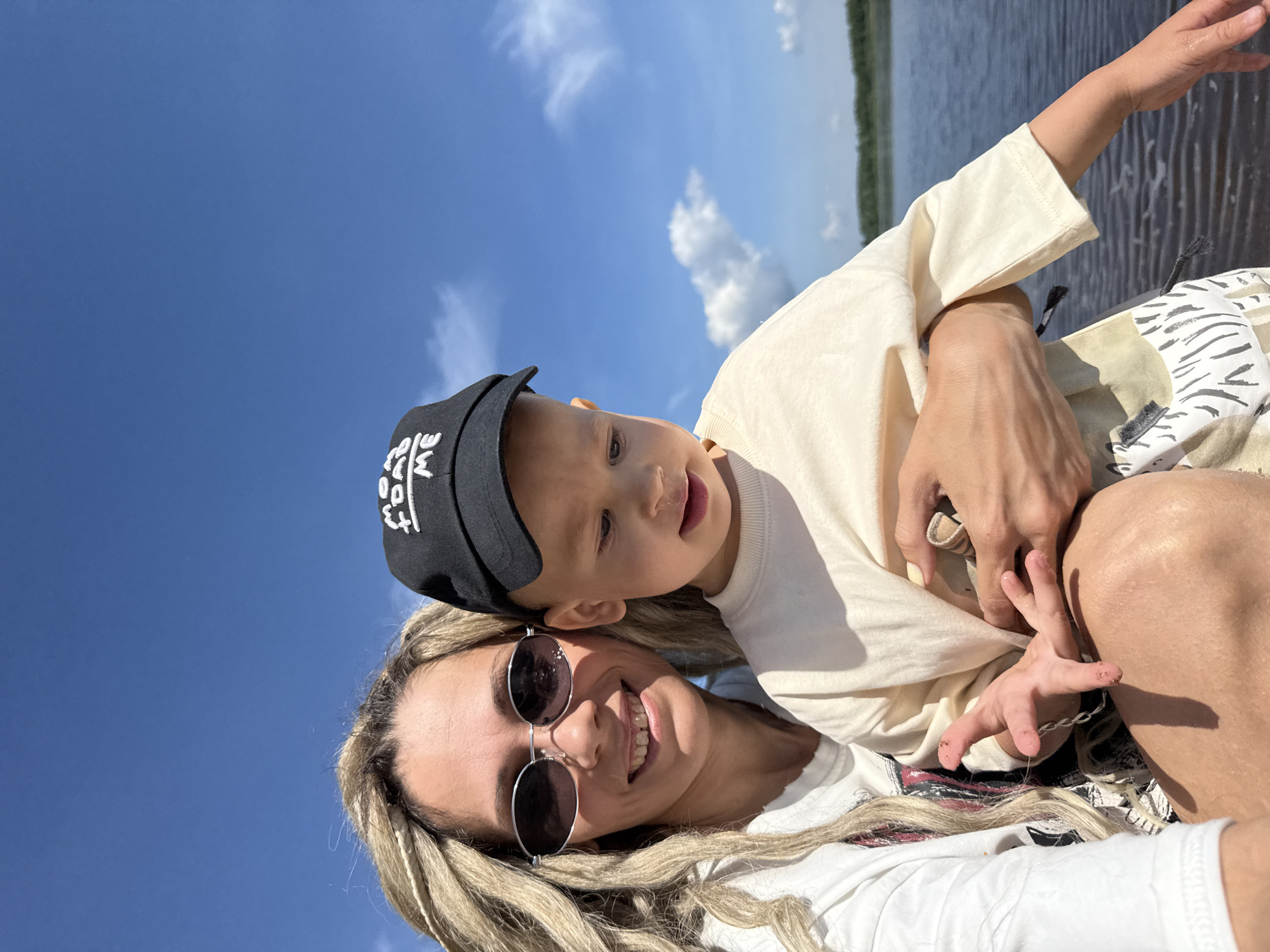 A mother with blonde dreadlocks and sunglasses is holding her young child on a sunny day near the water. The child is wearing a black baseball cap with white lettering and a light-colored t-shirt. The mother is wearing a white t-shirt. They appear to be on a boat or near a body of water, with a blue sky and clouds in the background. The location is identified as Rusanovo, Russia.