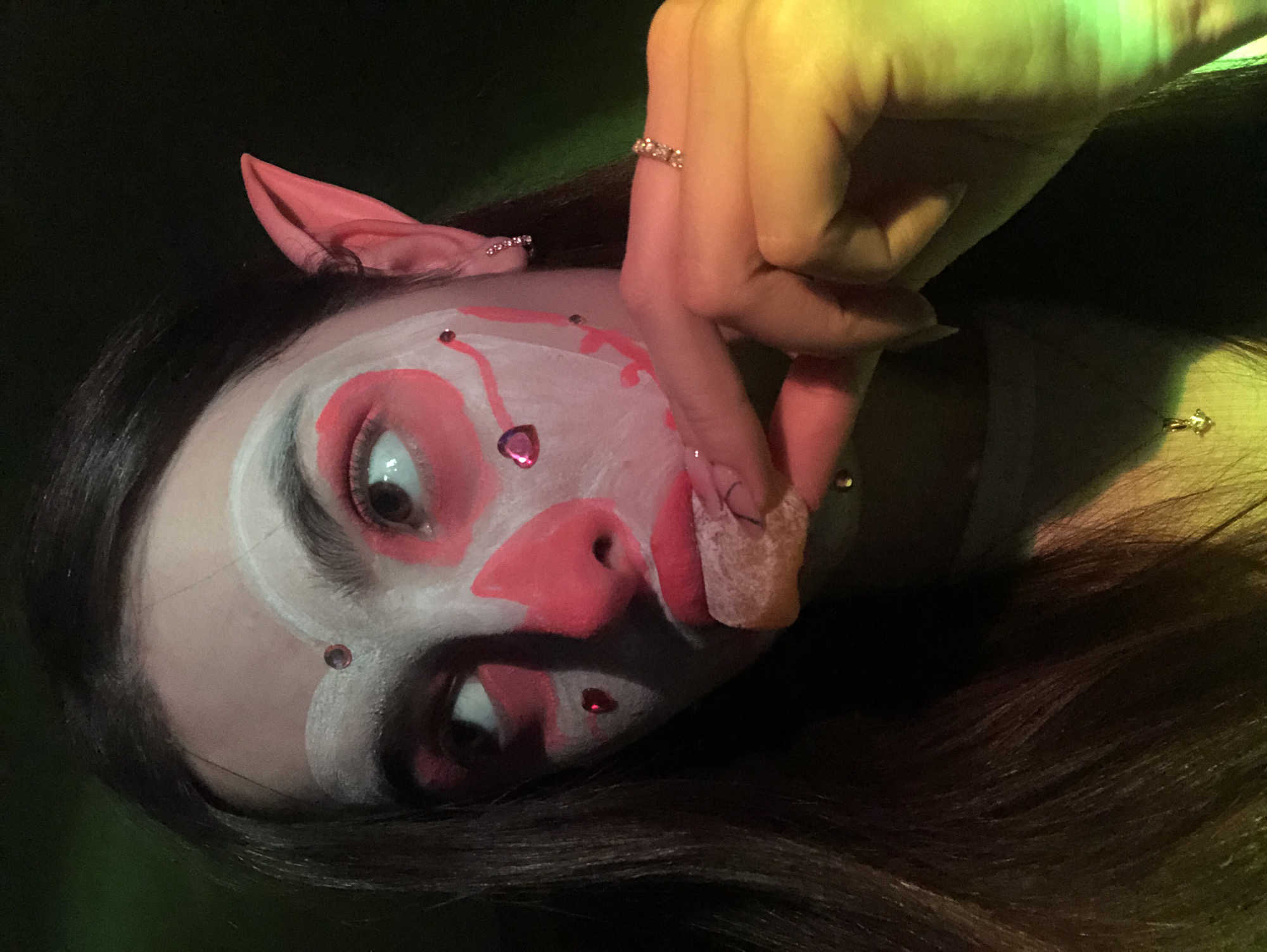 A person with clown makeup is lying on their side, wearing pink pig ears. The face is painted white with pink accents around the eyes, nose, and mouth. There are small, shiny gems on the face. A hand with a ring is near the face, touching the chin. The background is dark with green and yellow lighting.