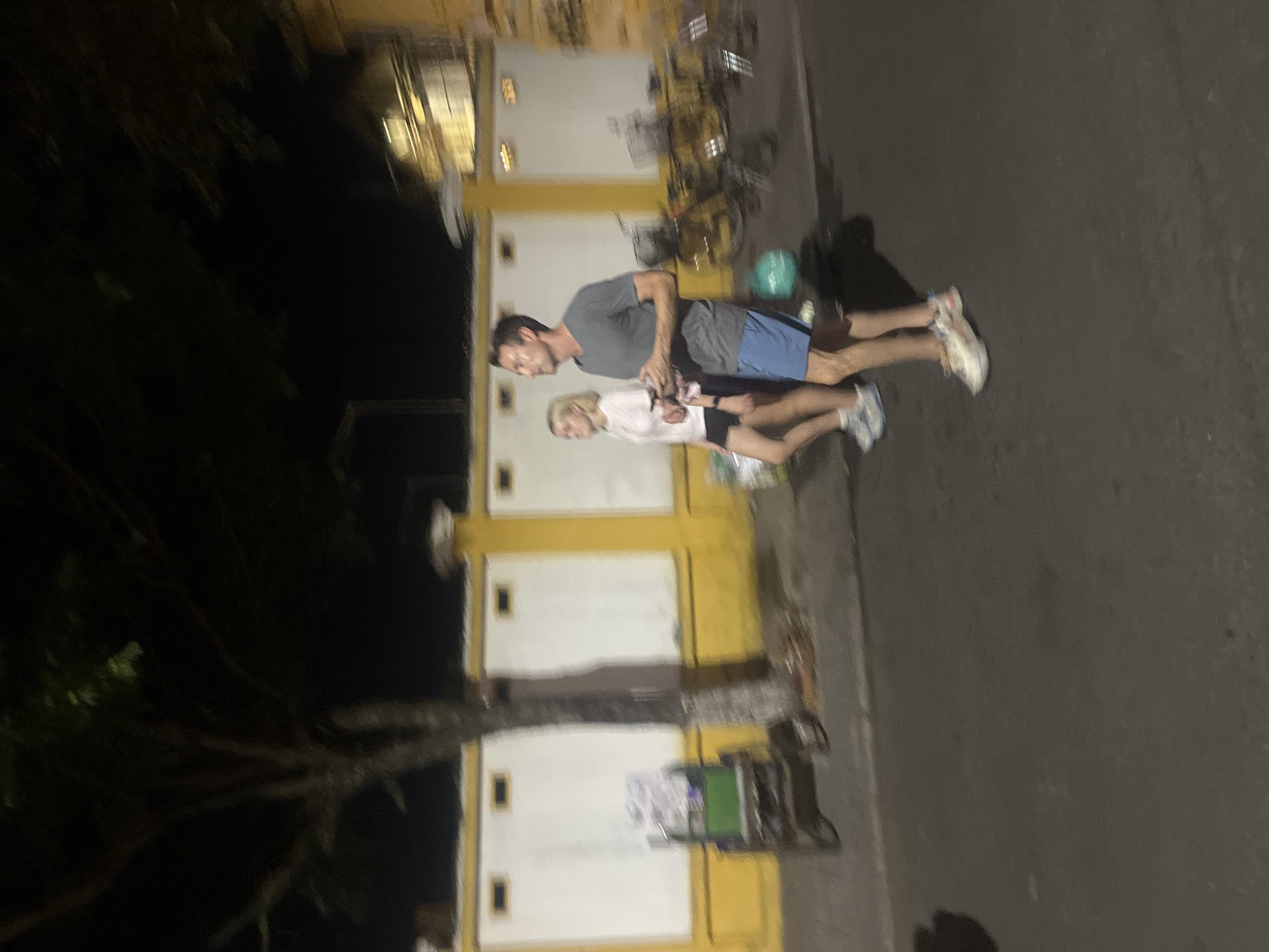 In Hội An Ward, Vietnam, two individuals walk along a street at night. The person on the right, a male, wears a gray t-shirt and blue shorts, with white and pink sneakers. He is looking to his left and holding a smartphone in his right hand. To his left, a female in a white t-shirt and dark shorts walks beside him, also looking forward and holding an object, possibly a phone, in her left hand. They are walking on an asphalt road in front of a yellow building with white panels and recessed rectangular shapes. A tree with visible branches stands to the left of the frame, next to a green cart with wheels. Bicycles with baskets are parked in the background, and the scene is illuminated by artificial light, creating shadows.