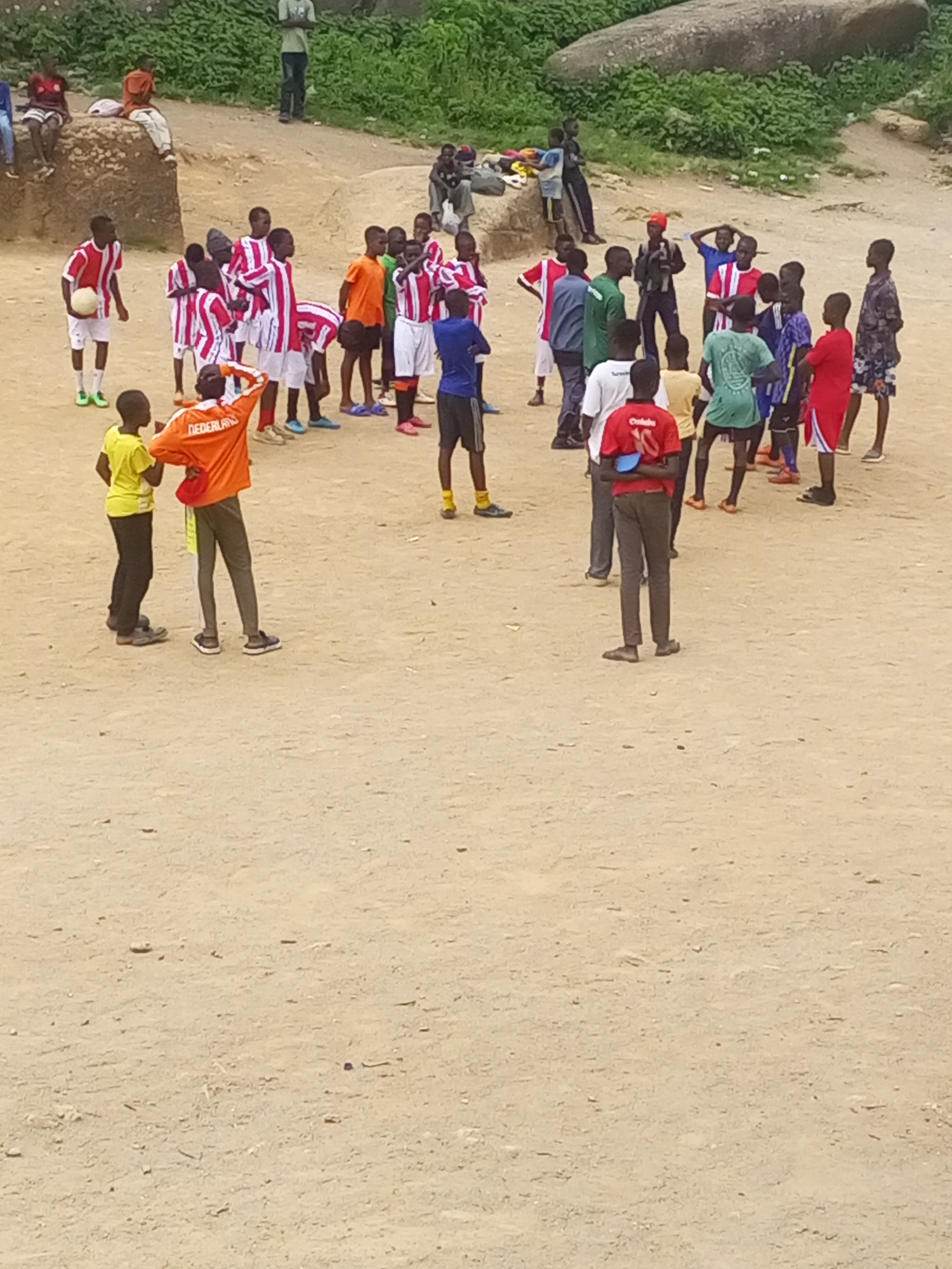 This image depicts an outdoor gathering of young males, likely a soccer practice, on a dusty, uneven ground in the city of Jos, Nigeria. The individuals, predominantly teenagers, are dressed in athletic attire, with many wearing red, white, and blue striped jerseys, indicative of a soccer team. A few are in plain colored shirts and shorts. Some are actively engaged, with one boy holding a soccer ball, while others are observing or conversing amongst themselves.
The environment is rural or semi-urban, characterized by a sandy, dirt ground, a large rock formation in the background, and sparse vegetation. The overall lighting suggests it is daytime, likely late morning or afternoon. The scene is populated by a group of approximately twenty individuals, all appearing to be male, and varying in age. The individuals are positioned at different distances from the camera, with some in the foreground and others further back in the mid-ground. The overall impression is one of casual activity and community engagement within a rudimentary outdoor setting.