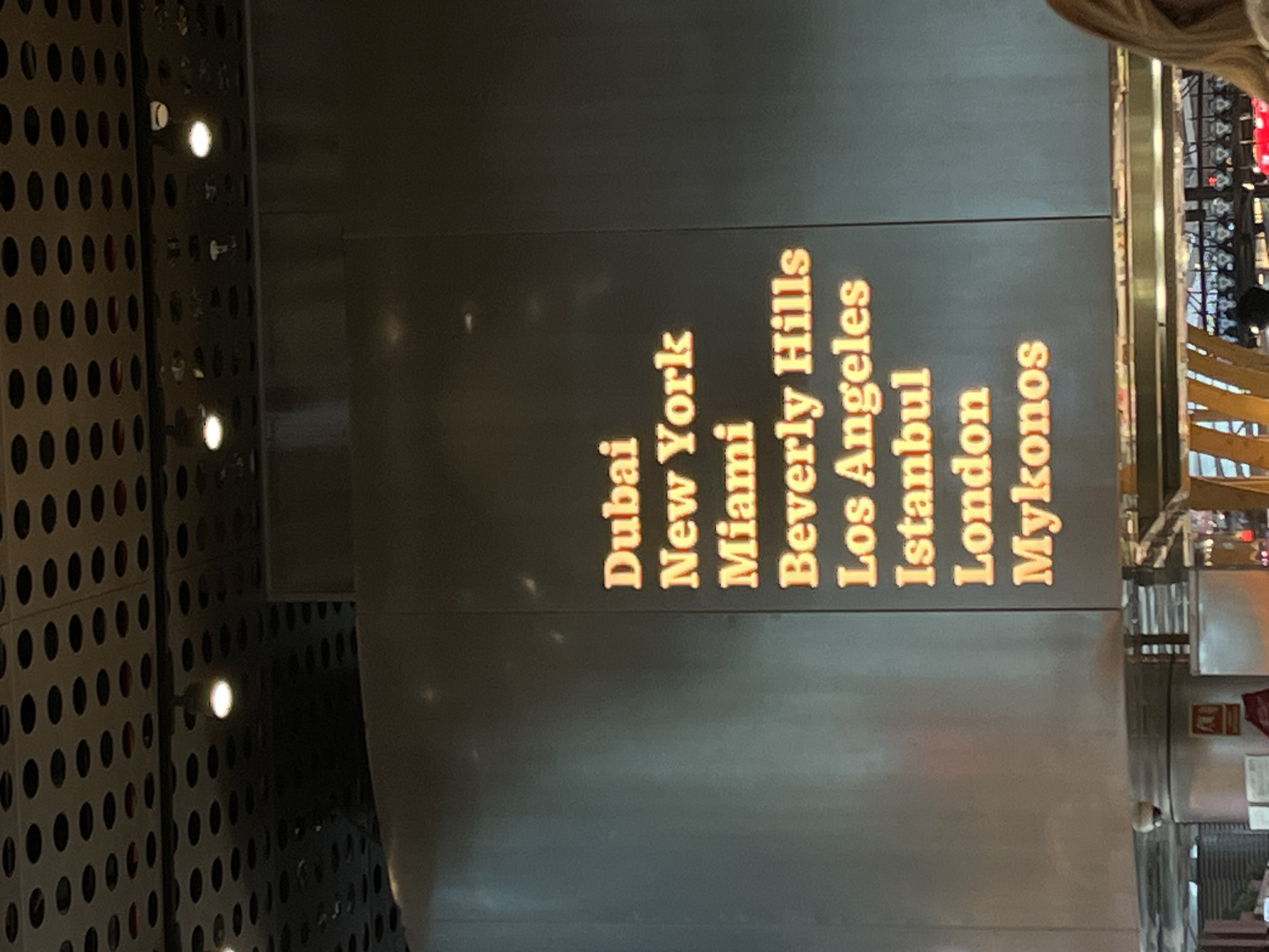 An interior photograph depicts a commercial establishment in an unknown city and location. The upper third of the image features a dark, perforated ceiling with numerous evenly spaced circular cutouts, through which multiple bright white light fixtures are visible. Dominating the mid-ground is a large, rectangular, dark gray panel displaying a vertical list of nine global cities in glowing orange-yellow serif font: "Dubai," "New York," "Miami," "Beverly Hills," "Los Angeles," "Istanbul," "London," and "Mykonos." The panel's surface exhibits subtle reflections. Below the panel, a horizontal metallic structure spans the width of the frame. Visible below this structure are elements suggesting a service or bar area, including a cylindrical column with a white dome surveillance camera mounted near its top, a red fire safety sign, and rows of bottles or glassware. The lower right portion of the image reveals a partial view of an individual's light-colored hair. The scene is illuminated by artificial light sources from the ceiling and the display panel.