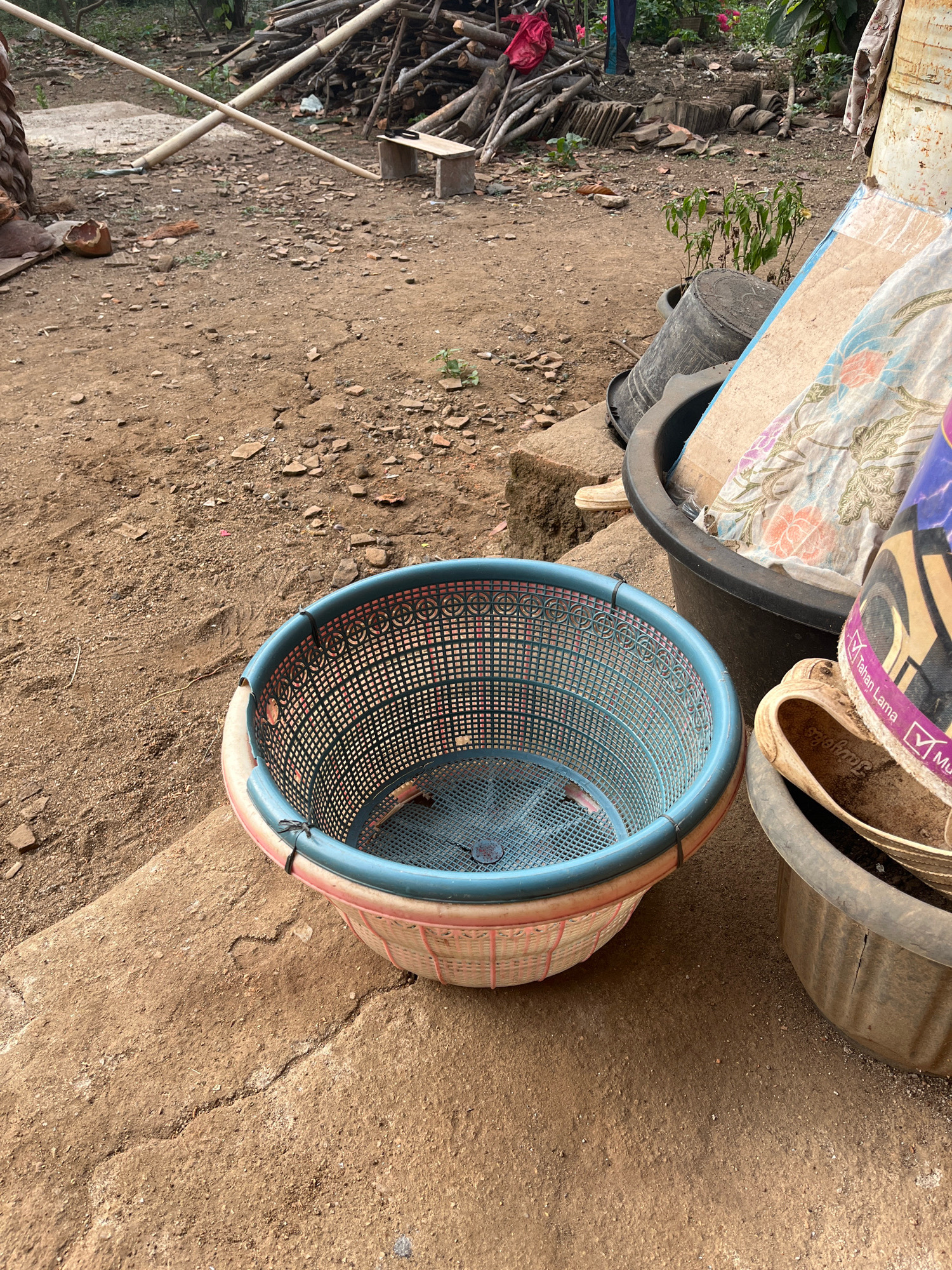 The image depicts an outdoor scene in Kebumen, Indonesia. In the foreground, a blue and pink plastic basket with a grid pattern sits on a concrete surface. To the right of the basket, a large black plastic tub contains fabric with floral patterns and other indeterminate items. Further to the right, a grey plastic tub holds a pair of worn, tan sandals. The ground is covered in dirt, small rocks, and scattered debris. In the background, a pile of chopped wood is visible, along with some bamboo poles. A small, green plant grows near the black tub. The overall environment appears to be rustic and unkempt.
