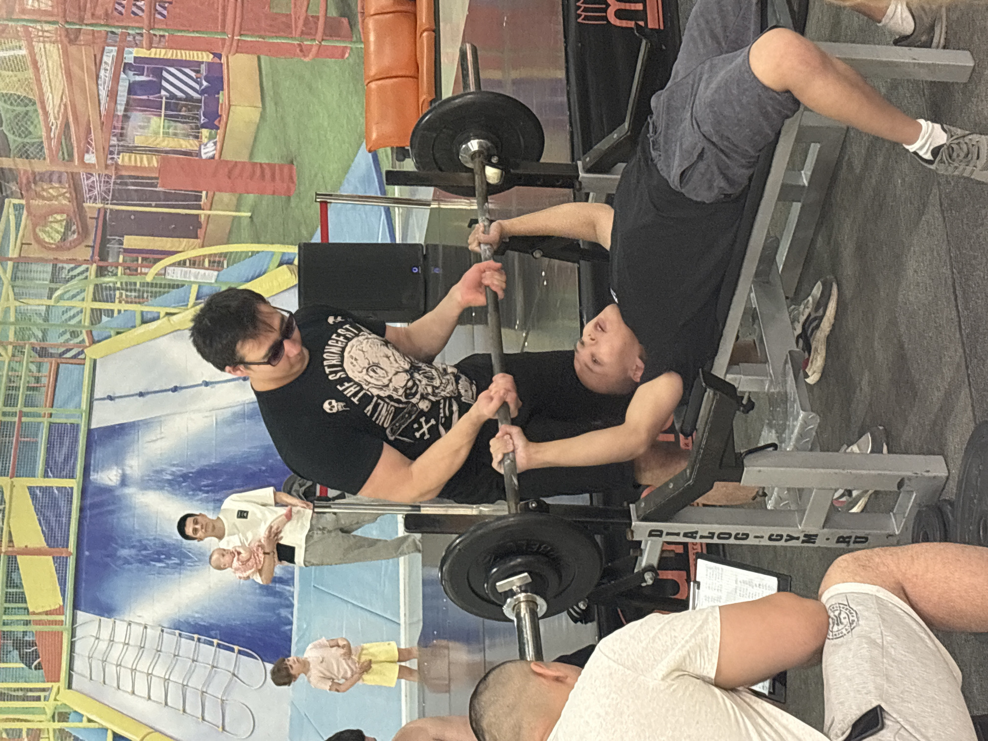 The image depicts an indoor scene that appears to be a gym or recreational facility, potentially located in Tri Potoka, Russia. In the foreground, a man is lying on a weight bench, performing a barbell bench press. Another man, wearing a black t-shirt with a skull graphic, is standing over him, spotting the lift and holding the barbell. To the left, a person in a white t-shirt and grey shorts is seated, facing away from the camera, with a clipboard and pen on a rack in front of them. In the background, a man is holding a baby, standing next to a young child. The environment features colorful, patterned walls, suggesting a play area or theme. A large blue slide structure is visible, along with orange seating in the distance. The floor is dark and appears to be rubberized, common in gyms. There are also various pieces of gym equipment and metal racks present.