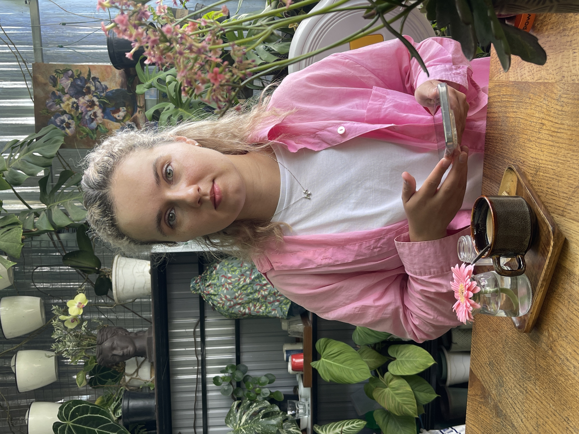 The image shows a woman in a pink shirt and white t-shirt seated at a wooden table. She is holding a mobile phone. On the table is a dark-colored coffee cup on a wooden tray and a small vase with a pink flower. The background features a wall with plants in white pots. Also visible is a painting of flowers. The location is Da Lat, Vietnam.