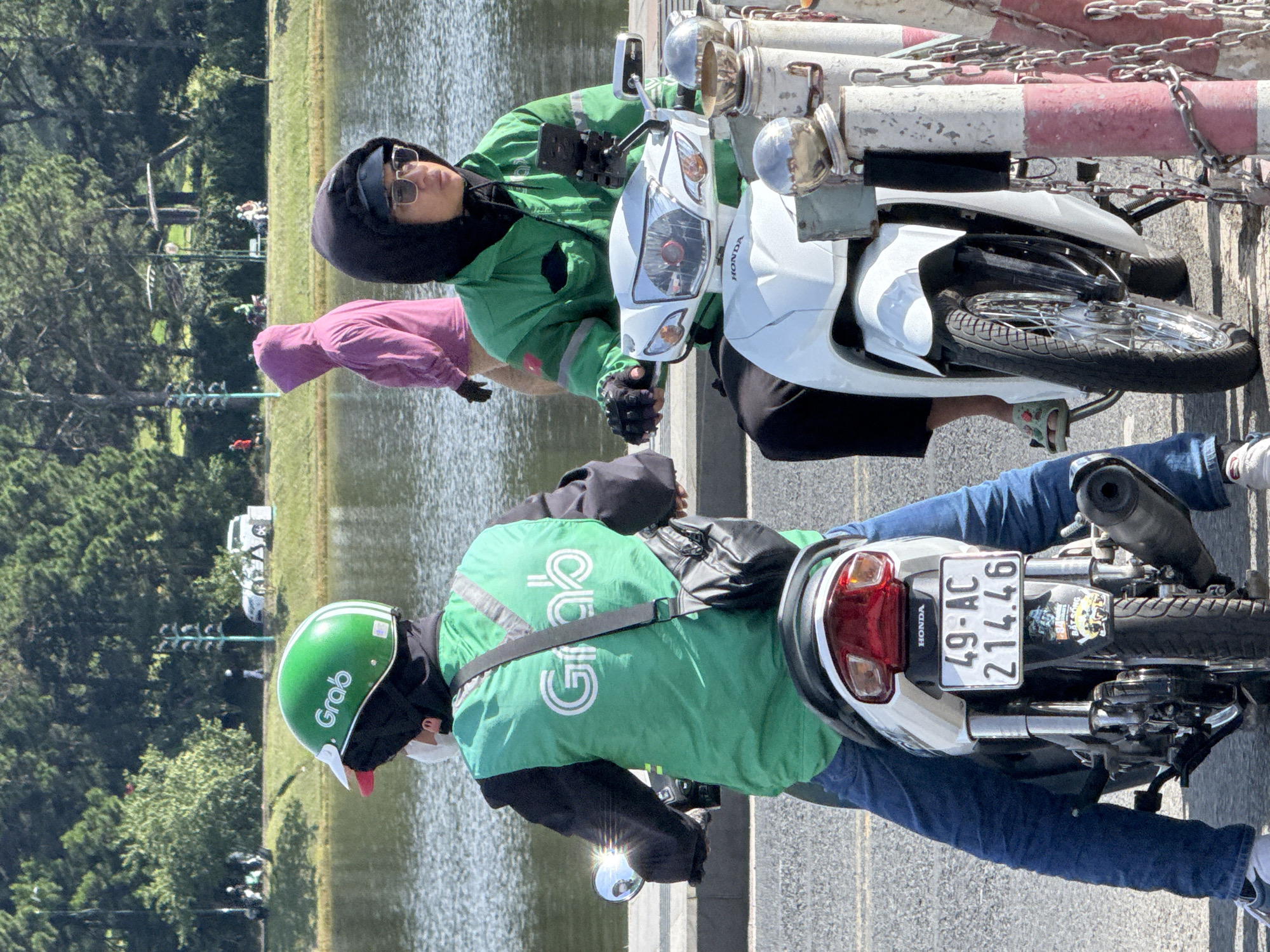 Two individuals wearing green Grab uniforms are seated on scooters. The scooter on the left has a license plate reading "49-AC 214.46". The scooter on the right has a phone mounted on its handlebars. In the background, a person in a pink hooded jacket walks near a body of water, with trees and a distant building visible. The scene appears to be on the promenade of Xuan Huong Lake in Da Lat, Vietnam, characterized by the presence of the lake, surrounding trees, and paved walkway.