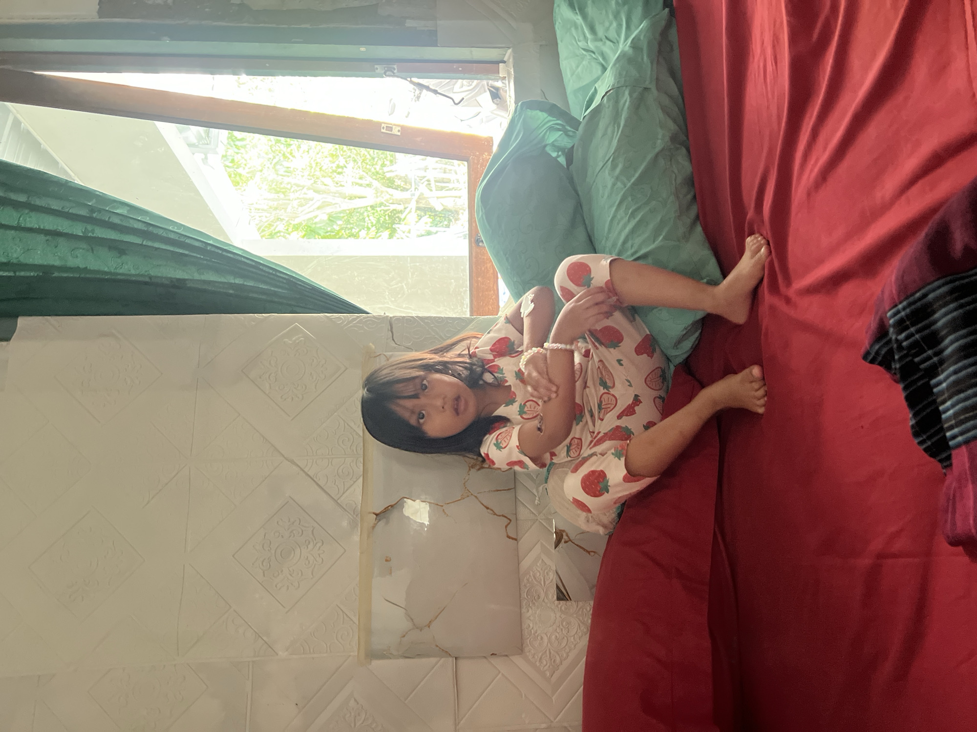 A young girl with dark hair and eyes, wearing pajamas with a strawberry pattern, sits cross-armed on a bed with red sheets. She is positioned against a textured wall with diamond-shaped patterns. Two teal pillows are visible behind her. To the right, a green curtain is drawn back to reveal an open window with bright sunlight streaming in, illuminating trees and foliage outside. The scene appears to be an indoor bedroom in a residential area of Kecamatan Pati, Indonesia.