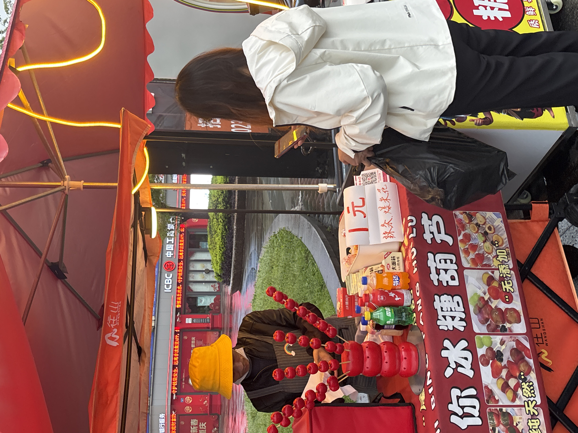 The image depicts a street food scene in Jiangjin, China. A woman in a white jacket and black pants is standing at a food stall, looking at her phone. She is holding a black plastic bag. The stall vendor, wearing a yellow hat, is preparing sugar-coated haws (tanghulu). Various snacks and drinks are visible on the stall. Signage includes Chinese characters for the ICBC bank, "Wangshan" (the brand name), and promotional text for the tanghulu. A street and a building facade are visible in the background.