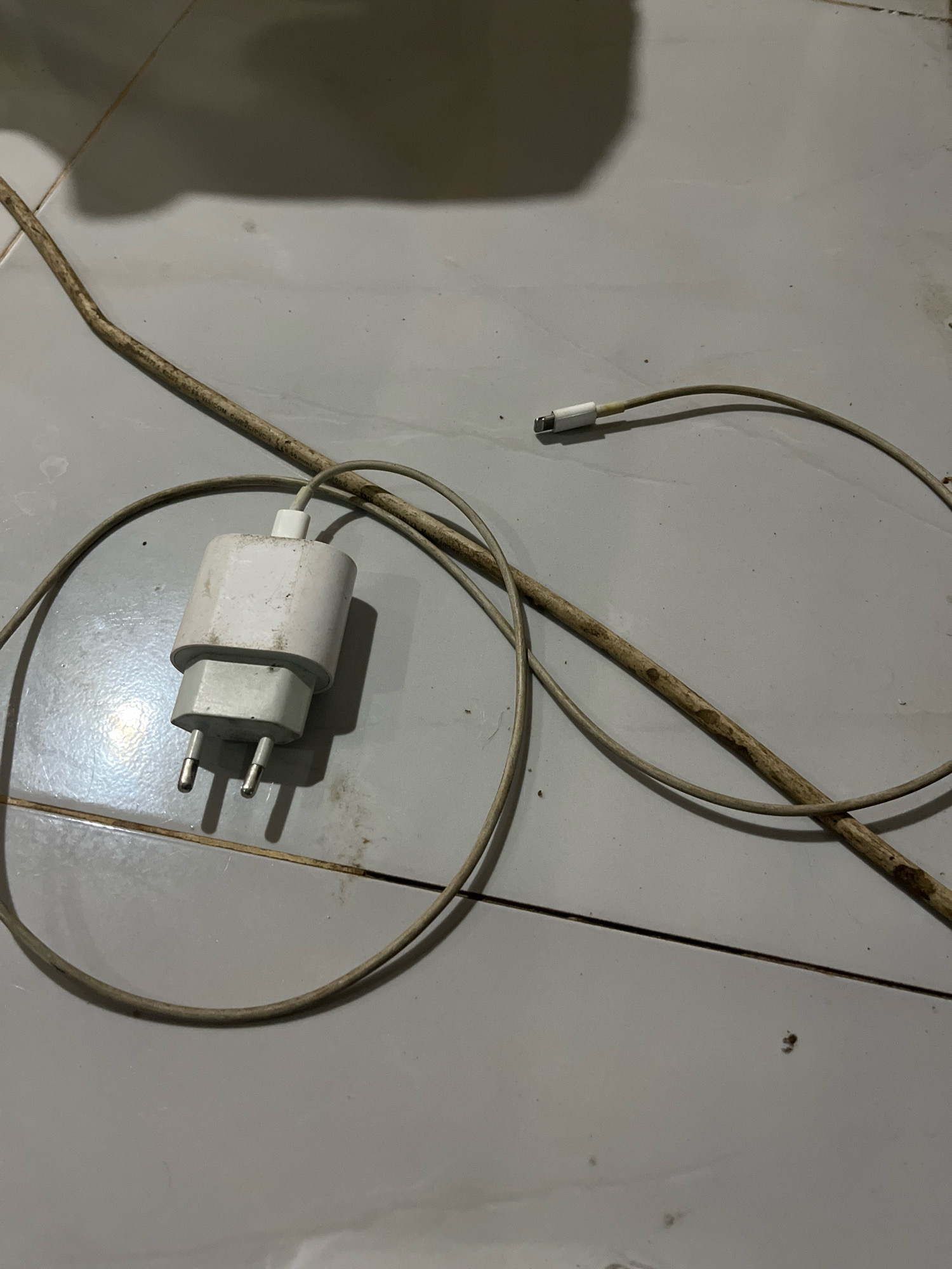 The image depicts a white power adapter with two European-style prongs, connected to a light gray charging cable. The cable extends and splits, with one end featuring a metallic connector, likely a USB-C or Lightning port. A thin, dirty brown stick lies across the cable. The objects are situated on a light gray tiled floor, with visible grout lines. The scene appears to be indoors, possibly in a residential setting within Margodadi, Indonesia, given the context provided. There are no people visible in the image, and the focus is solely on the discarded charging accessories and the stick. The lighting suggests artificial illumination, casting shadows on the floor.